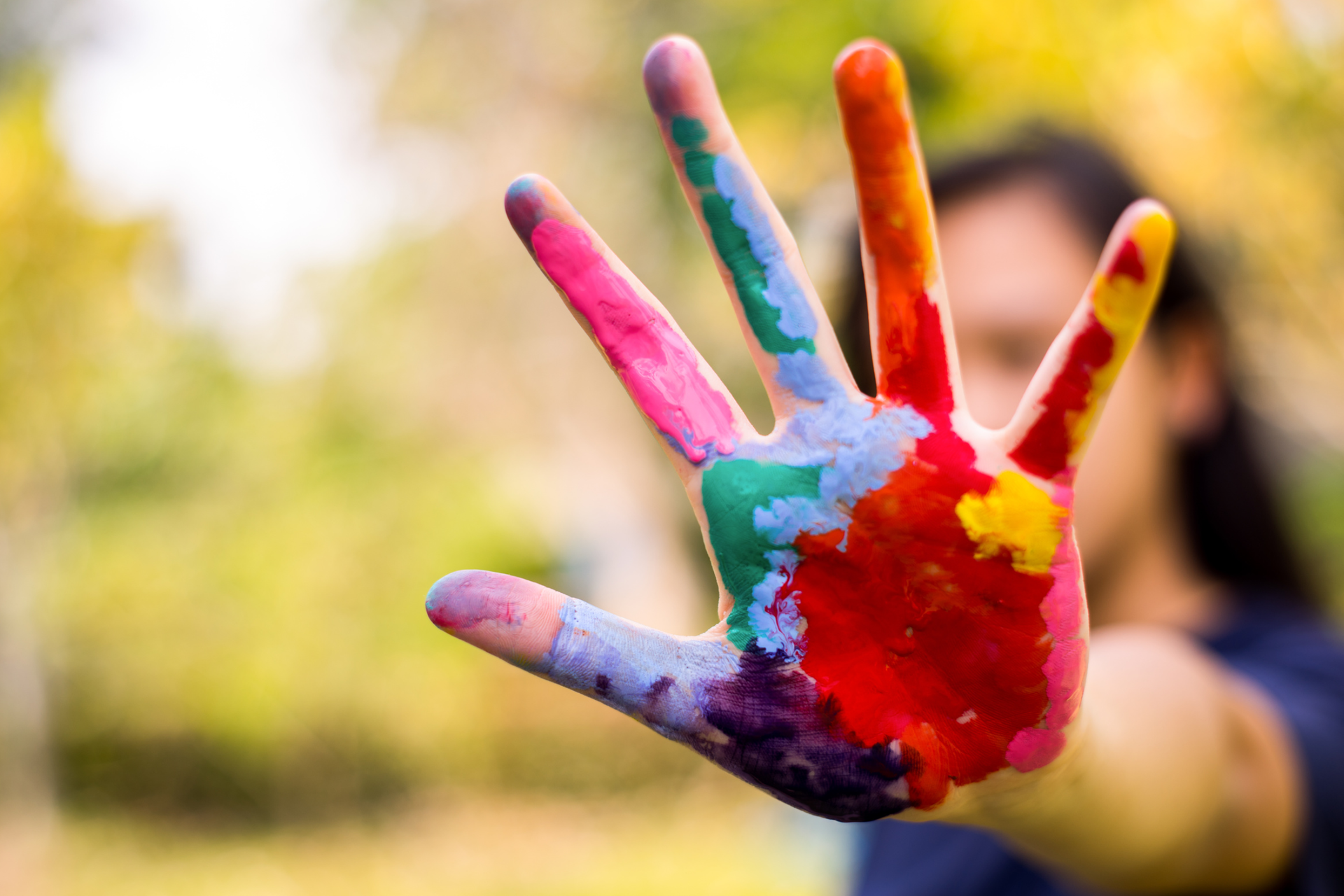 Ilustracja przedstawia fotografię wyciągniętej dłoni, otwartej wewnętrzną stroną w kierunku widza. Cała dłoń jest pomalowana grubą warstwą kolorowych farb. Dłoń znajduje się na pierwszym planie, tło jest rozmyte.