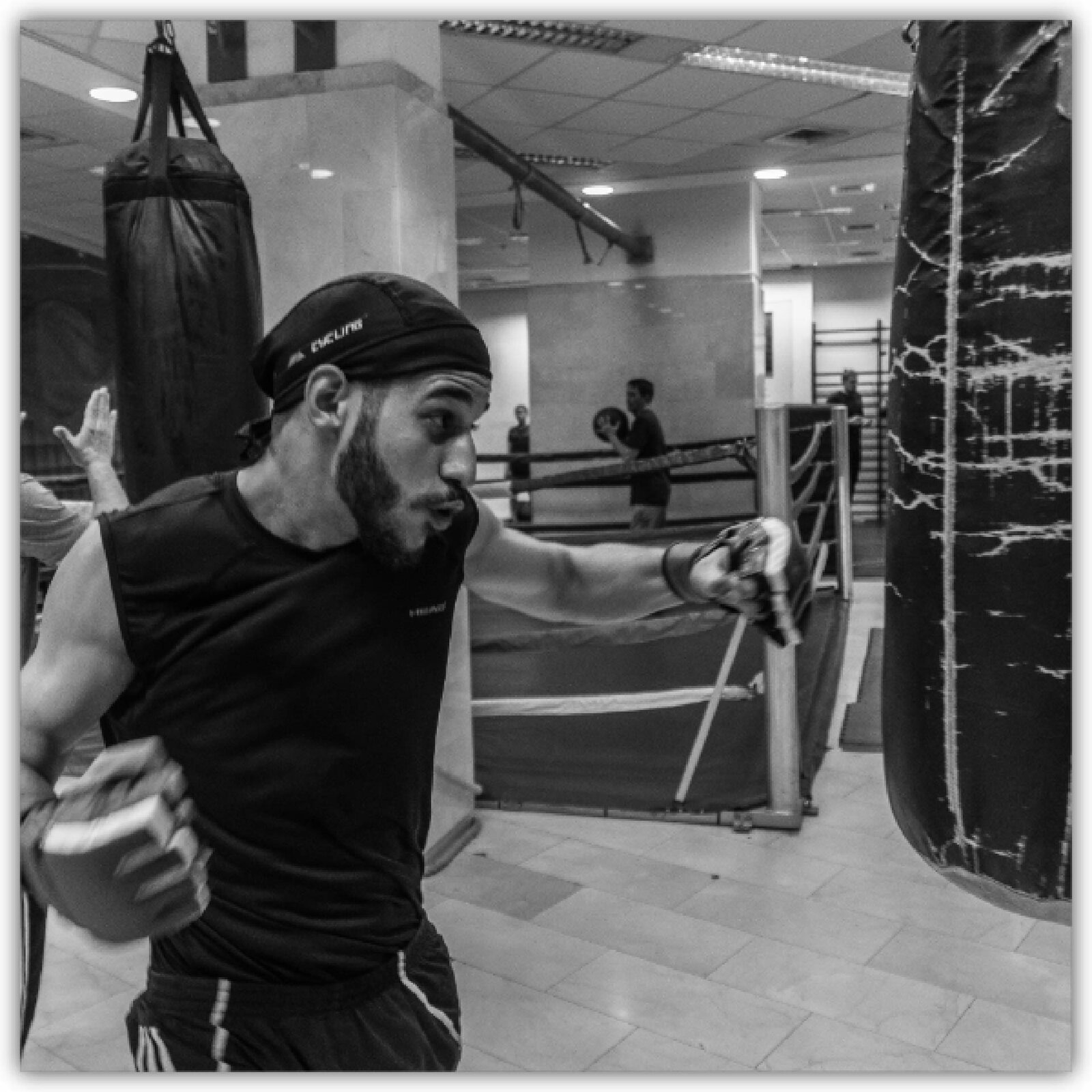 Ilustracja przedstawia mężczyznę boksującego worek. W tle ring oraz drugi worek przy którym ktoś ćwiczy.