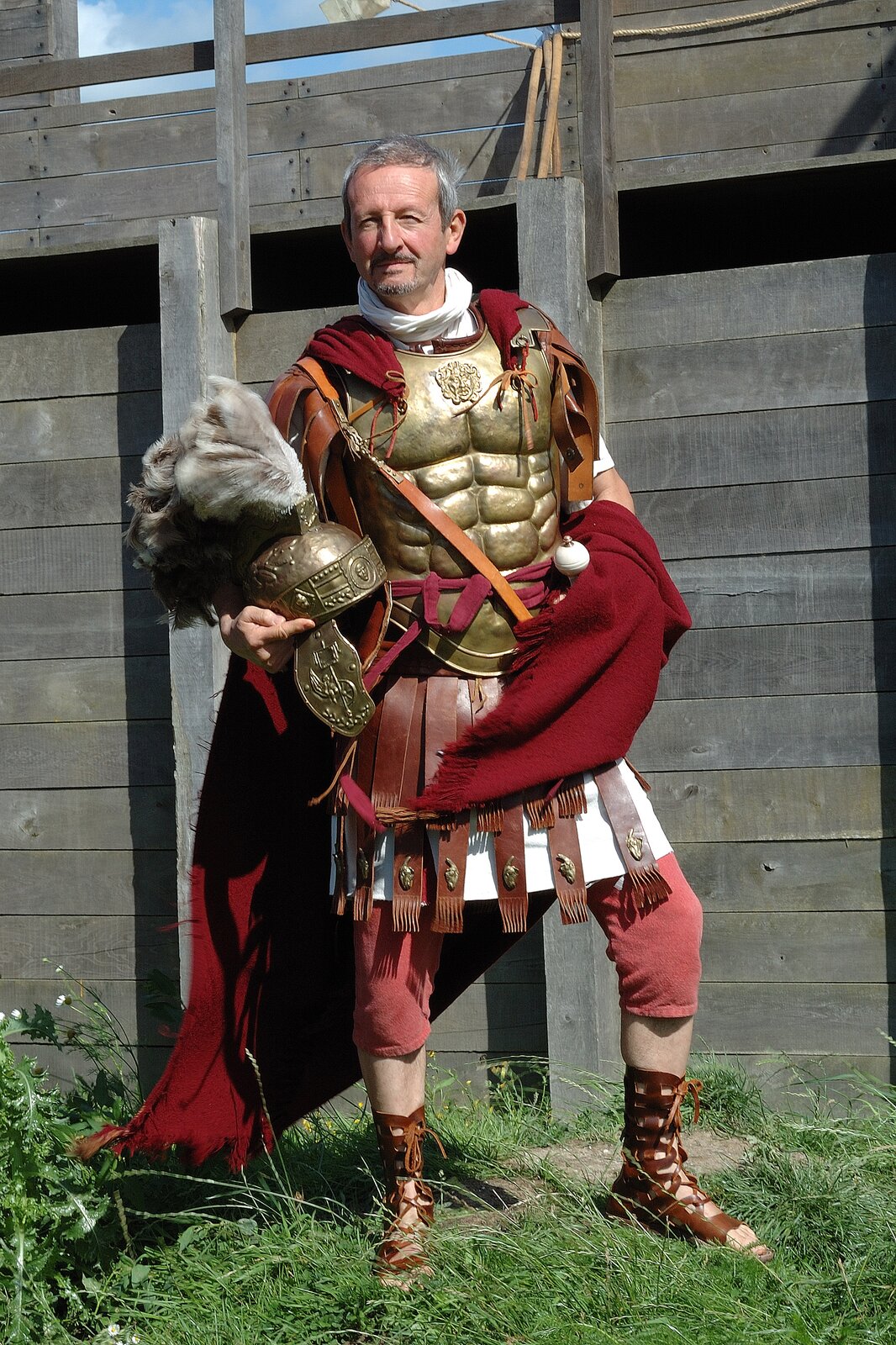 Fotografia ukazuje mężczyznę prezentującego replikę zbroi rzymskiego trybuna wojskowego. Jest to mężczyzna w średnim wieku o siwych włosach, krótkiej brodzie oraz wąsach. Mężczyzna mruży oczy – słońce oświetla mu twarz. Stoi on na tle szaro‑zielonej drewnianej konstrukcji. Ma na sobie czerwone spodnie sięgająca za kolano, brązowe wiązane sandały, czerwoną pelerynę, złotą zbroję oraz złoty hełm z szarym pióropuszem, trzymany w prawej ręce. Mężczyzna stoi na trawie. 