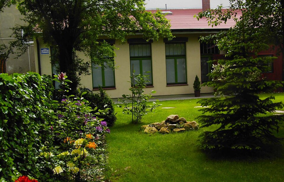 Zdjęcie przedstawia parterowy budynek z wysokimi oknami i spadzistym dachem. Elewacja jest jasnożółta, okna są w zielonych ramach. Przed budynkiem jest ogród z trawnikiem, kwiatami oraz drzewami.