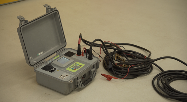 Ilustracja przedstawia Miernik MI 3252 MicroOhm 100A. Urządzenie jest wbudowane w szarą plastikową skrzynkę z uchwytem. Do miernika podpięte są cztery kable. Urządzenie wyposażone jest w panel sterujący oraz mały ekranik wyświetlający informację. 