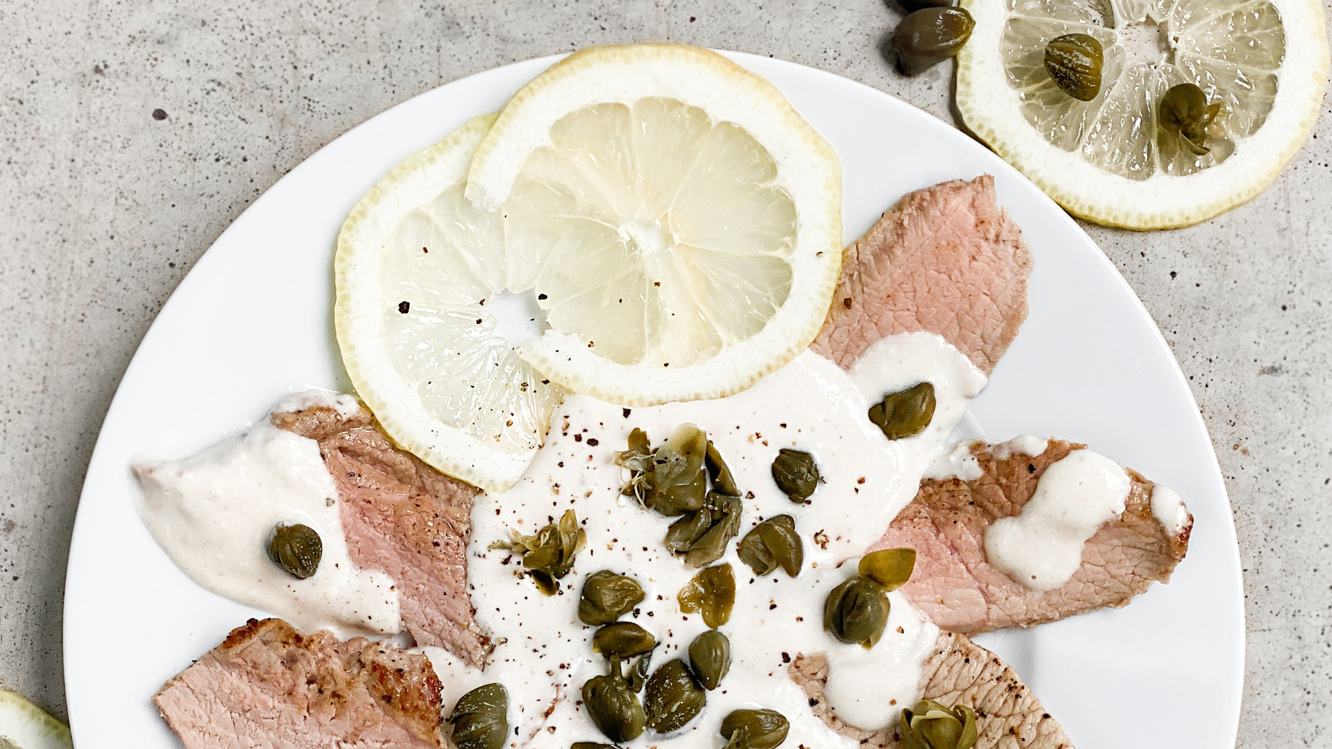 Zdjęcie wykonane z góry przedstawia włoskie danie Vitello tonnato. Na talerzu znajdują się małe, cienko pokrojone plasterki mięsa. Mięso polane jest jasnym sosem. Całość posypana jest kaparami. Na talerzu znajdują się również dwa plasterki cytryny. Obok talerza na blacie leży plasterek cytryny, a na nim kapary.