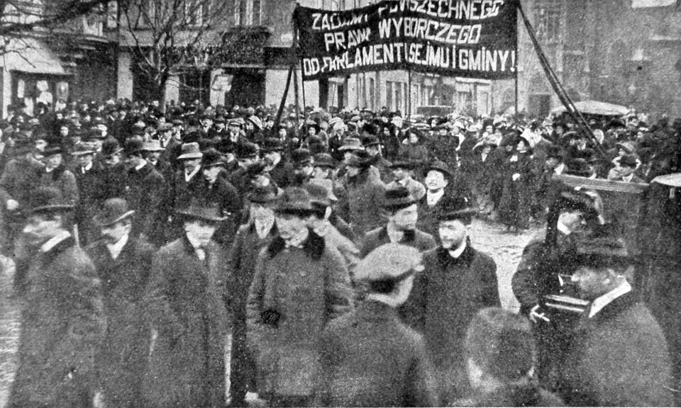 Czarno‑białe zdjęcie przedstawia tłum mężczyzn i kobiet na ulicy miasta. Kobiety protestują. Kilka z nich trzyma duży transparent z napisem: Żądamy powszechnego prawa wyborczego do parlamentu, sejmu i gminy. Na ulicy, na pierwszym planie stoi duża grupa mężczyzn w płaszczach, na głowach mają meloniki i kapelusze. W tle kamienice, niewielkie drzewa oraz samochody.