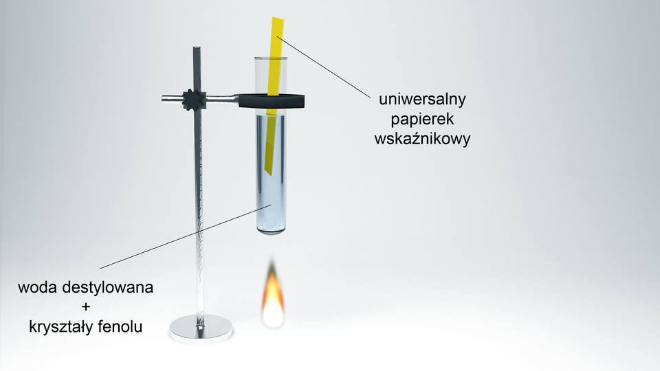 Ilustracja przedstawiająca statyw laboratoryjny z metalową łapą, w której umieszczono probówkę. W probówce znajduje się woda destylowana oraz kryształy fenolu. Pod probówką znajduje się płomień palnika. Do probówki włożono uniwersalny papierek wskaźnikowy. 