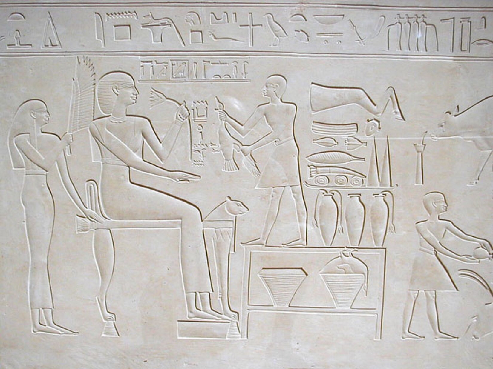 Na reliefie w białym kamieniu przedstawiono kilka postaci. Największa postać siedzi na krześle i trzyma w jednej ręce kwiat. Mniejsza postać, stojąca za nią, trzyma, skierowany w stronę większej postaci wachlarz z dużego liścia palmowego. Najmniejsza postać, która stoi na stole z drugiej strony największej postaci, trzyma w obu rękach gęś. Ponadto, na reliefie przedstawiono wazy, hieroglify, liście.