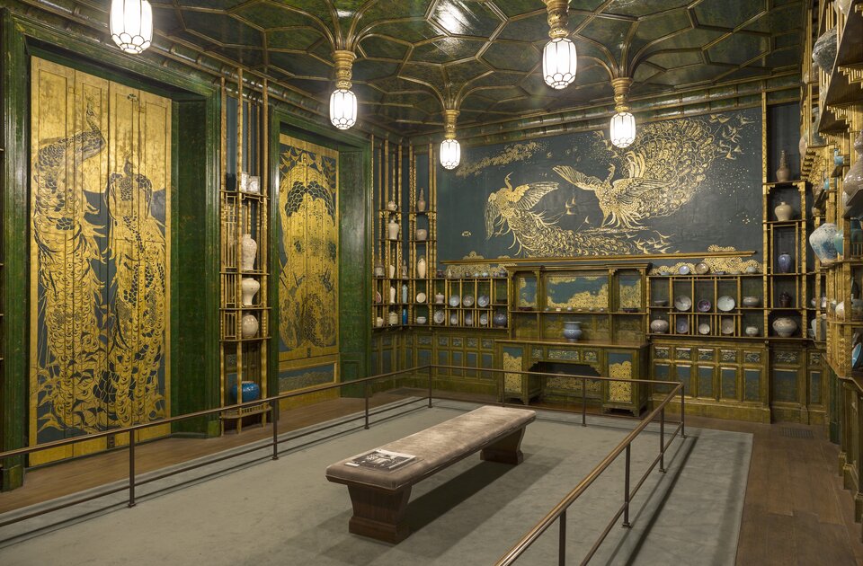 Na zdjęciu widoczne są ściany po lewej stronie i naprzeciwko widza. Pośrodku stoi na szarym dywanie drewniana, obita materiałem ława, na której końcu leży gazeta. Ławę otacza metalowa balustrada. Na czarnej ścianie naprzeciwko są namalowane złote pawie. Przy nich stoją złote, wąskie, wysokie półki, na których są ustawione ozdobne wazy. Na ścianie po lewej stronie znajdują się zielone futryny, w których są umieszczone złoto‑czarne parawany, jeden z nich jest ozdobiony złotymi, dużymi pawiami.  Natomiast na drugim parawanie  jest umieszczona złota kobieta ubrana w kimono. Przy ścianie naprzeciwko stoi antyczny, złoty kredens. Niebieskie fronty są ozdobione złotymi motywami kwiatowymi. Na środku stoi porcelanowy wazon. Powyżej na półce są ustawione bardzo małe naczynia. Obok kredensu po dwóch stronach znajdują się półki, na których stoją wazy. Nad nimi znajduje się złota listwa. Przy suficie w formie sklepienia kolebkowego wiszą białe żyrandole. Oświetlają one pomieszczenie.