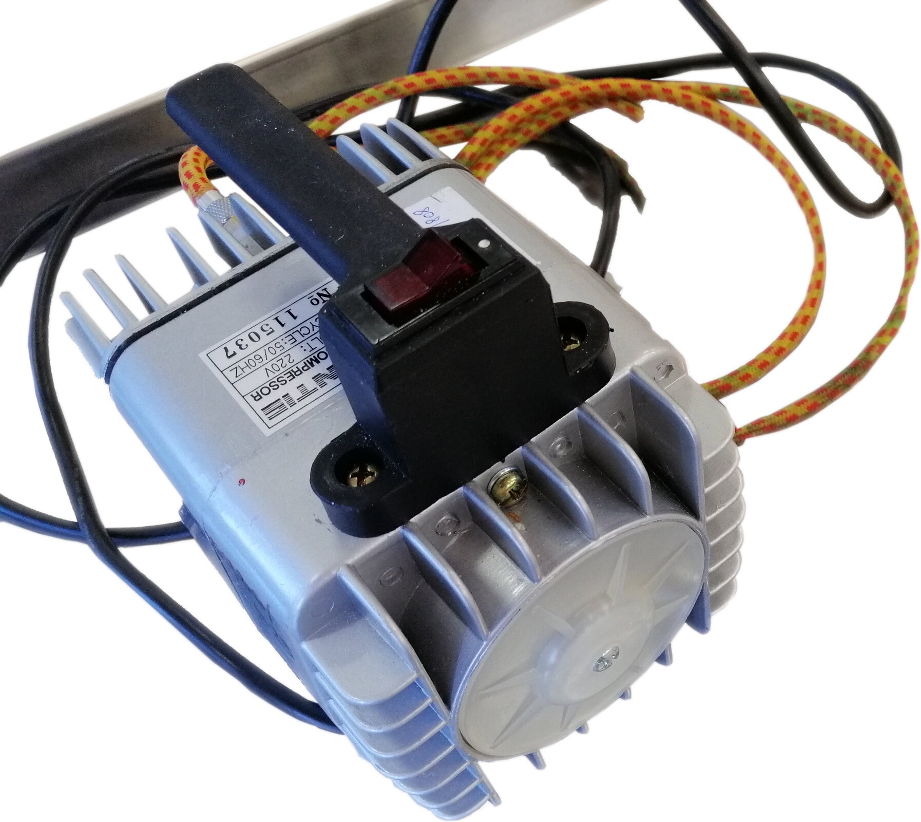 Na zdjęciu widoczny jest kompresor. Widoczny jest prostopadłościenny srebrny pojemnik, na górze którego przymocowany jest czarny uchwyt, od którego odchodzą kable w różnym kolorze.