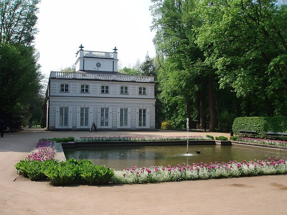 Zdjęcie przedstawia wygląd Białego Domku. Jest to drewniana, tynkowana budowla na planie kwadratu, ozdobiona na krawędzi dachu balustradą oraz belwederkiem w środku. Okna budynku są prostokątne i wysokie. Okna na parterze posiadają otwarte okiennice. Elewacja jest zdobiona sztukaterią. Przed budynkiem widoczna jest sadzawka z fontanną. Wokół rosną krzewy i drzewa. 