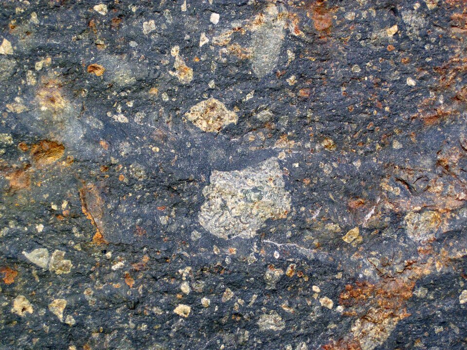 Na zdjęciu znajduje się czarna skała z wieloma jasnymi i rdzawymi wtrąceniami. 