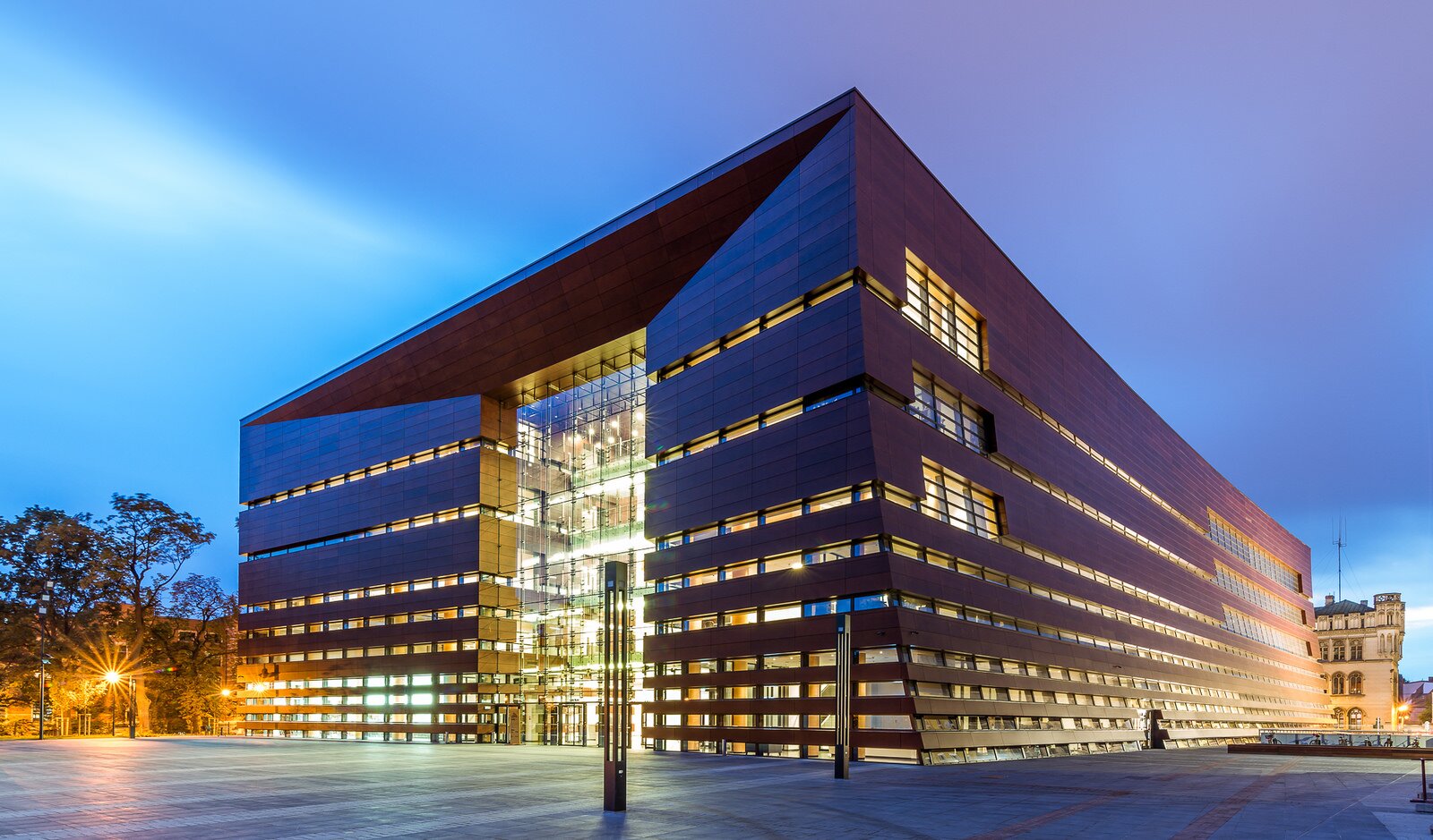 Fotografia przedstawia budynek Narodowego Forum Muzyki im. Witolda Lutosławskiego we Wrocławiu. Budynek ma kształt prostopadłościanu prostokątnego, z poziomymi liniami okien przypominających strun na gryfie gitary. Wejście przypomina otwór rezonansowy. Kształt obiektu oraz materiał imitujący drewno zastosowany na elewacji przypominają pudło rezonansowe instrumentu strunowego.