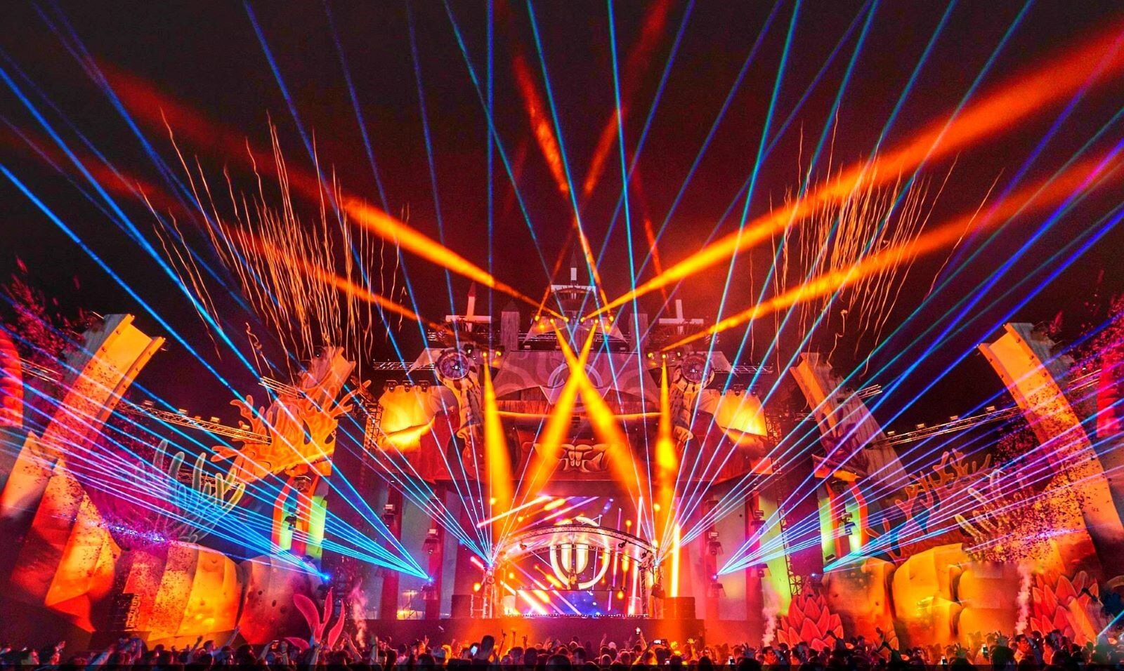 Zdjęcie pokazuje scenę koncertową w oprawie świateł. Wiązki silnych, cienkich promieni laserowych wychodzą z kilku źródeł od strony sceny i krzyżują się z szerokimi pasmami świateł reflektorów. Wszystkie światła i lasery w barwach ciepłych i zimnych przenikają się wzajemnie. Scena i całe jej otoczenie stało się jednym, pulsującym w rytm muzyki obrazem. W dole tłum tańczących ludzi z podniesionymi rękami.