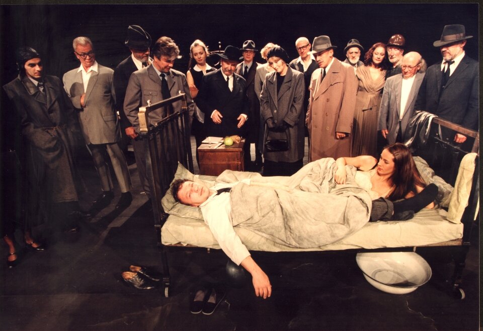 Zdjęcie ze spektaklu "Kartoteka". W centrum znajduje się metalowe łóżko. Leżą w nim dwie postaci: mężczyzna ubrany w koszulę i spodnie ma głowę po lewej stronie łóżka. Kobieta w białej koszuli nocnej ma głowę po prawej stronie łóżka. Są przykryci beżową kołdrą. Wokół łóżka stoją ludzie, którzy się im przyglądają. Wyglądają, jakby gdzieś przechodzili, ponieważ są ubrani w płaszcze, kurtki, czapki. 