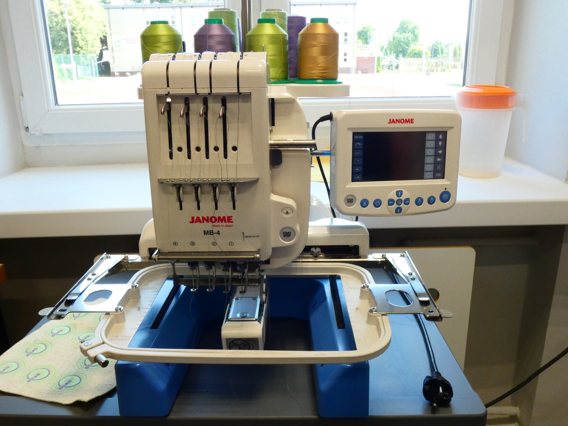 Zdjęcie przedstawia duże białe plastikowe urządzenie stojące na stole. Składa się z głównej części, mechanizmu haftującego, po prawej stronie ma niewielki ekran, a z przodu znajduje się pulpit, na którym kładzie się materiał. 