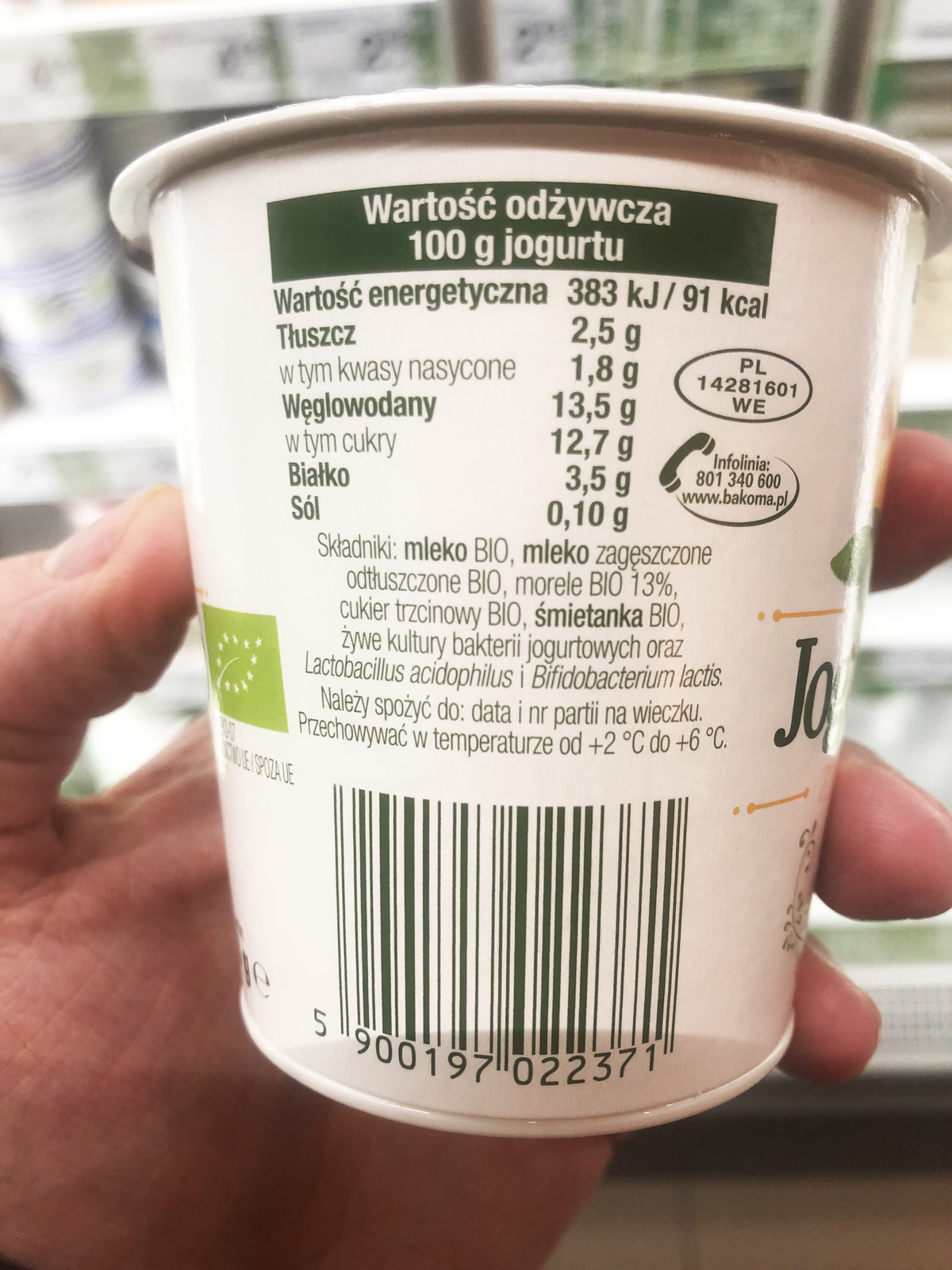 Ilustracja przedstawia etykietę jogurtu. Zawarto na niej następujące informacje: Wartość odżywcza na 100g jogurtu: Wartość energetyczna 383 kJ/ 91 kcal; Tłuszcz 2,5g w tym kwasy nasycone 1,8g; Węglowodany 13,5g w tym cukry 12,7g; Białko 3,5g; Sól 0,10g. Składniki: mleko BIO, mleko zagęszczone odtłuszczone BIO, morele BIO 13%, cukier trzcinowy BIO, śmietanka BIO, żywe kultury bakterii jogurtowych oraz Lactobacillus acisophilus i Bifidobacterium lactis. 