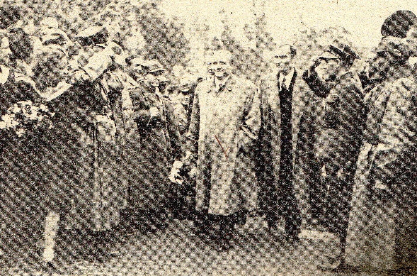 Zdjęcie przedstawia Stanisława Mikołajczyka, idącego w towarzystwie dwóch mężczyzn między tłumem ludzi złożonym z cywilów i wojskowych. Żołnierze salutują. Kobiety trzymają w dłoniach bukiety kwiatów.
