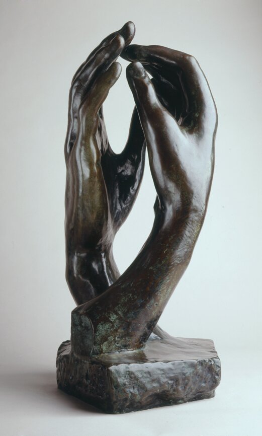 Zdjęcie przedstawia rzeźbę. To dwie uniesione ku górze dłonie. Jedna dłoń jest kobieca, druga męska. Stykają się palcami.