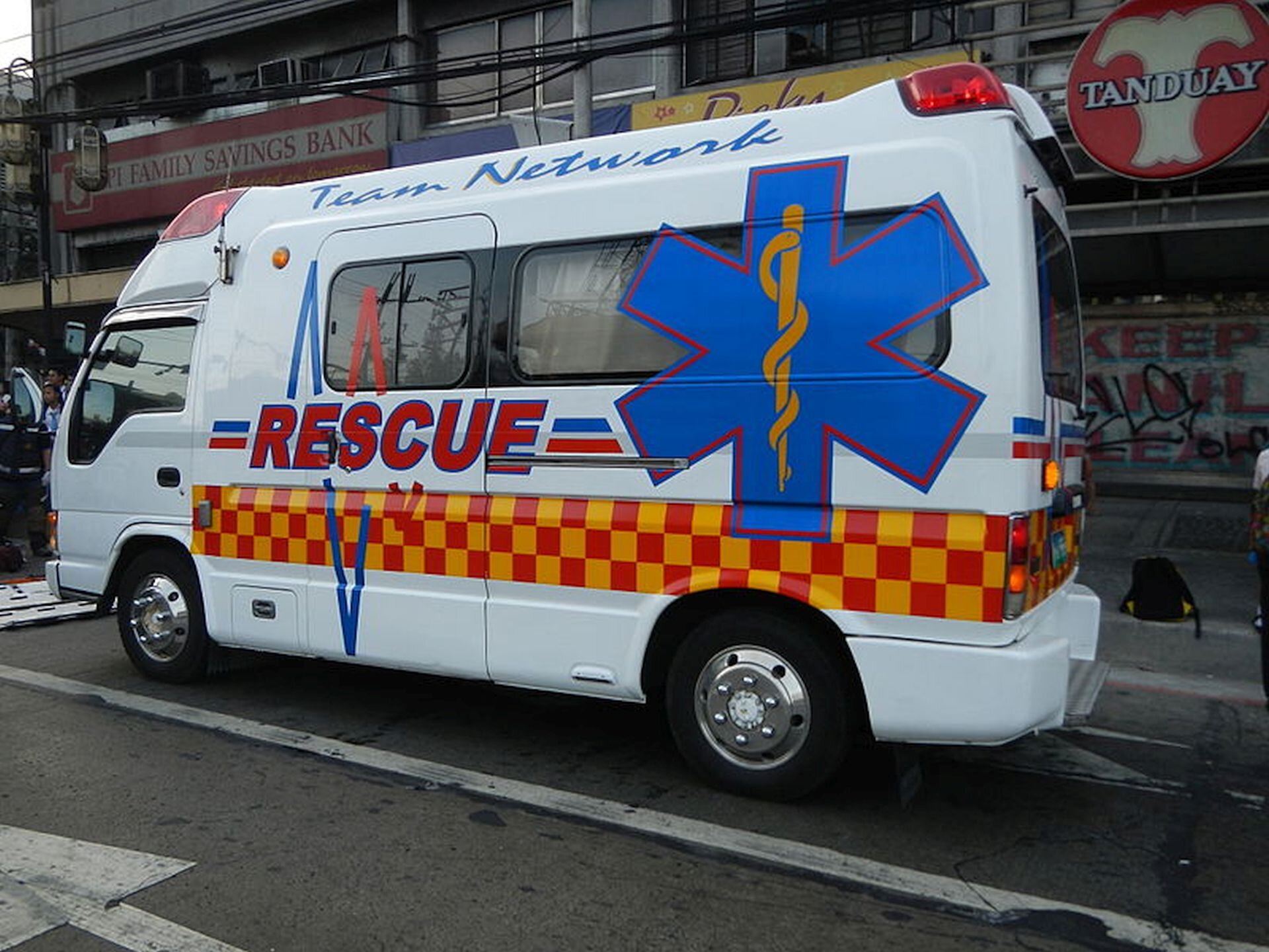 Ilustracja przedstawia angielski ambulans. Jest to biały samochód z dużą gwiazdą życia namalowaną na boku pojazdu wraz z napisem Rescue. Pojazd stoi na ulicy przed budynkami handlowymi.