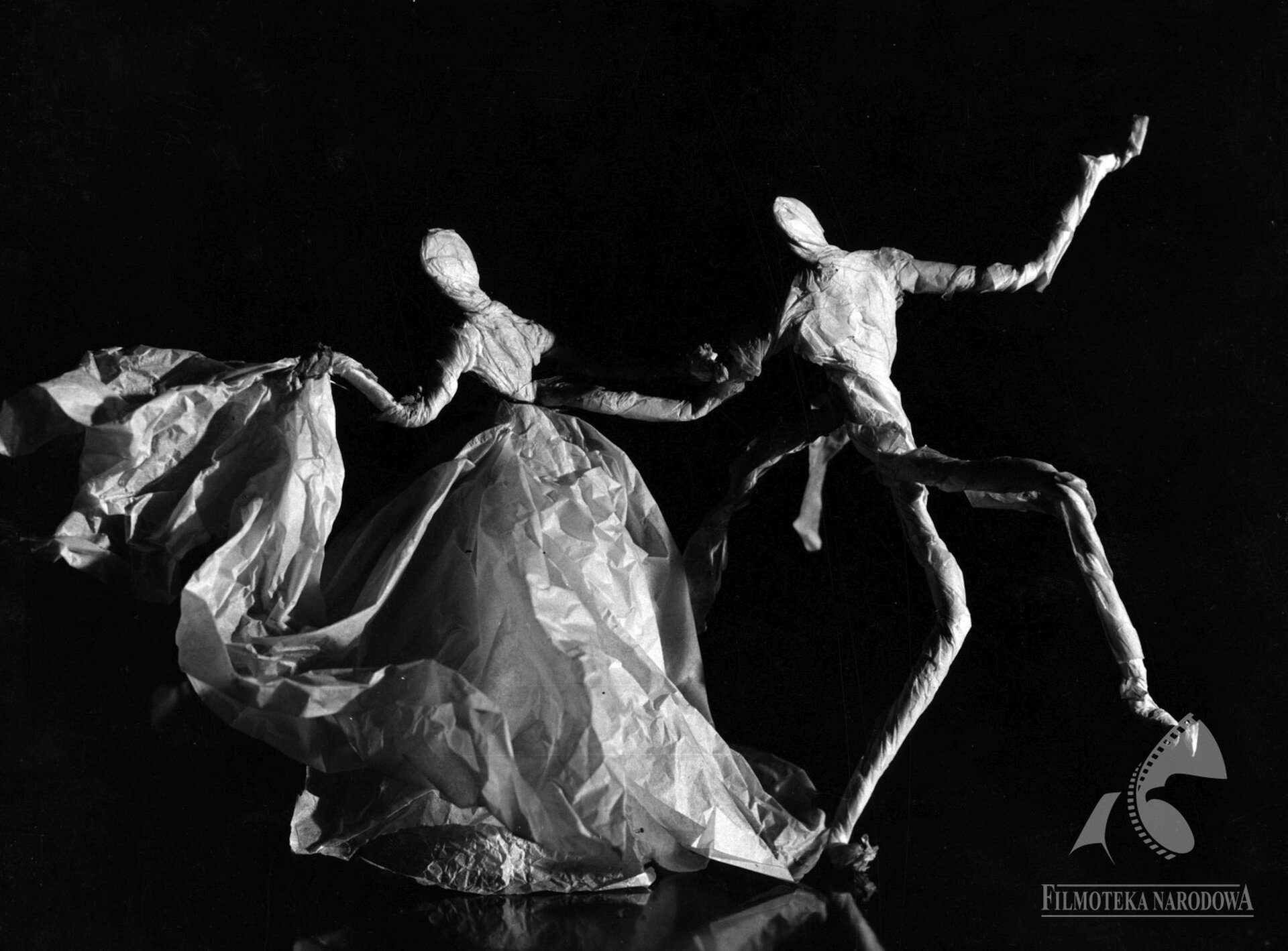 Ilustracja przedstawia kadr z filmu Witolda Giersza i Ludwika Perskiego „Oczekiwanie”. Ukazuje dwie tańczące postacie, wykonane z papieru. Para trzyma się za ręce, wykonuje gesty. Mężczyzna ma uniesioną jedną nogę i pochyla się w stronę kobiety, która w jednej ręce trzyma długą suknie. Mężczyzna ma założony frak.