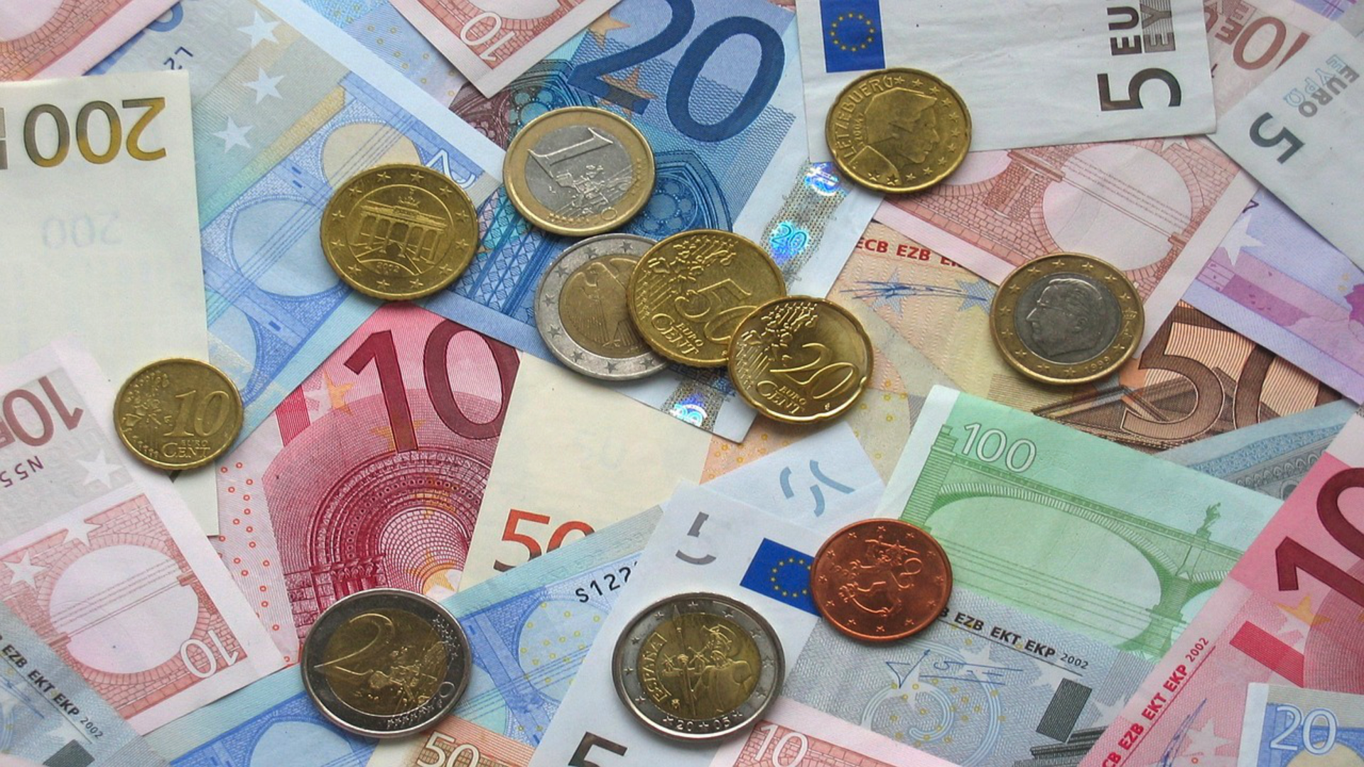 Zdjęcie przedstawia rozrzucone banknoty euro o nominałach: 5, 10, 20, 50, 100 i 200. Na banknotach leżą monety euro: 1, 2 euro oraz centy o nominałach: 10, 20, 50.