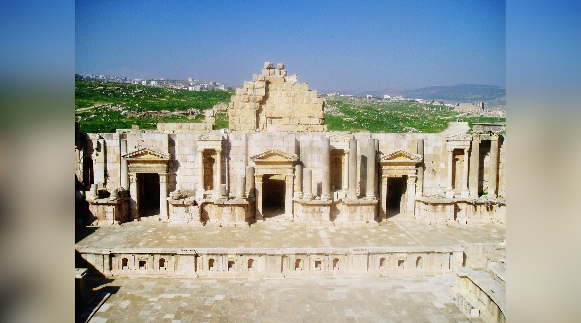 Zdjęcie przedstawia starożytną budowlę teatralną. Ukazana jest kamienna scena przypominająca budynek z trzema wejściami.