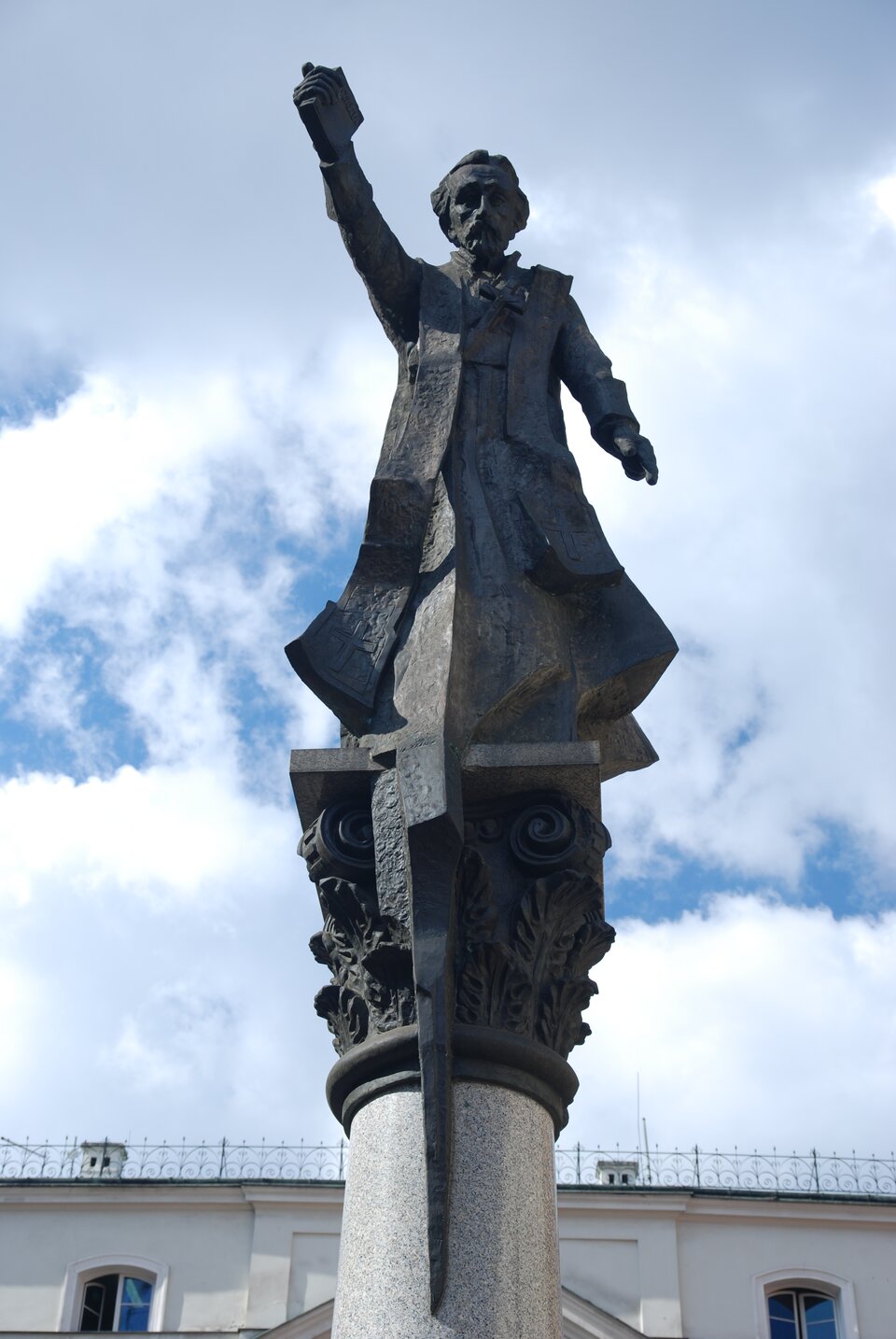 Zdjęcie przedstawia pomnik. Na owalnej kolumnie znajduje się postać stojącego mężczyzny. Jest w długiej szacie, ma przewieszoną przez szyję stułę. Trzyma uniesioną w górę prawą rękę, a w dłoni książkę.