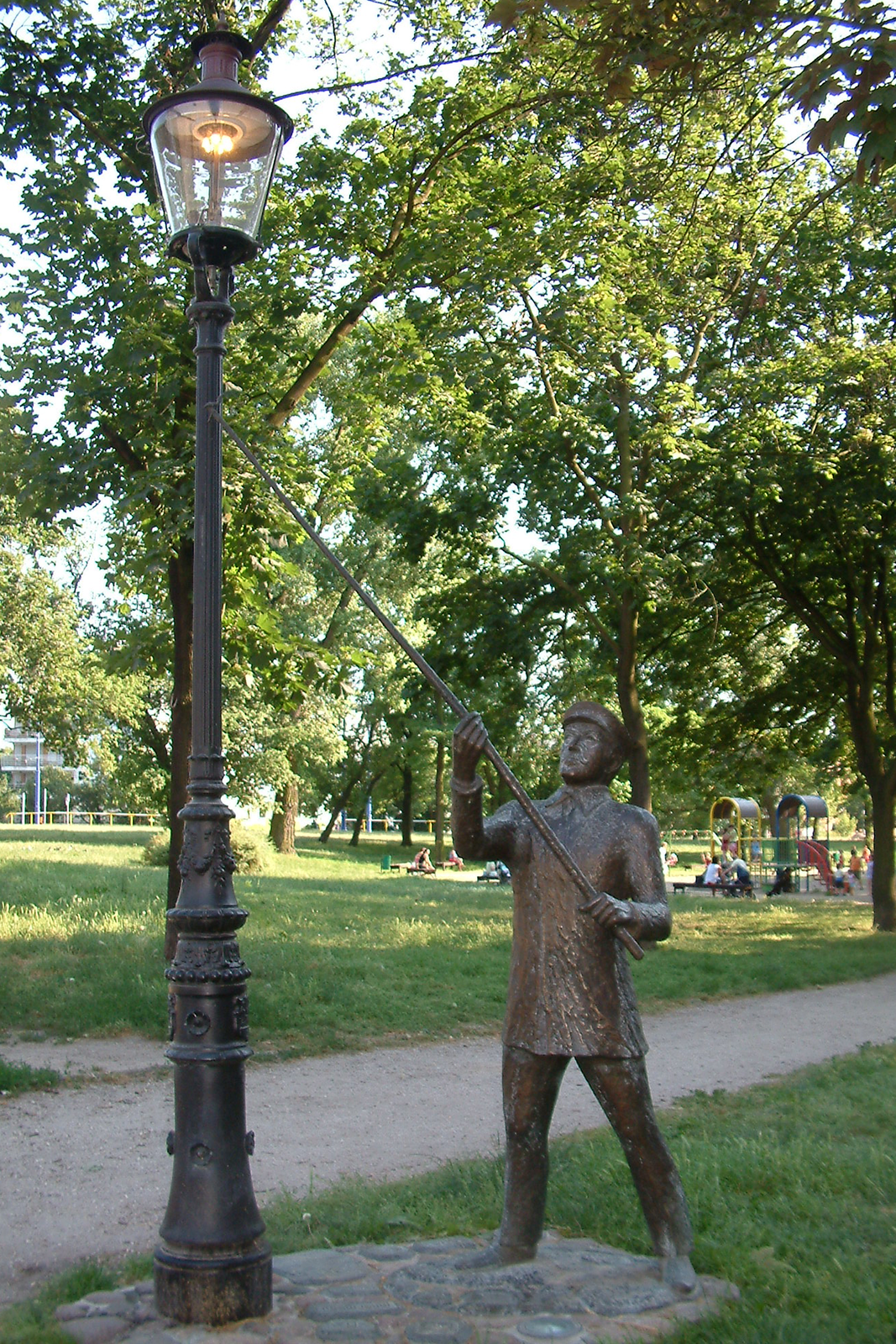 Na zdjęciu widoczna jest rzeźba latarnika w parku. Jest ona postawiona obok latarni. Latarnik trzyma w rękach długi kij z knotem na czubku, który służy do zapalania płomienia w latarni.