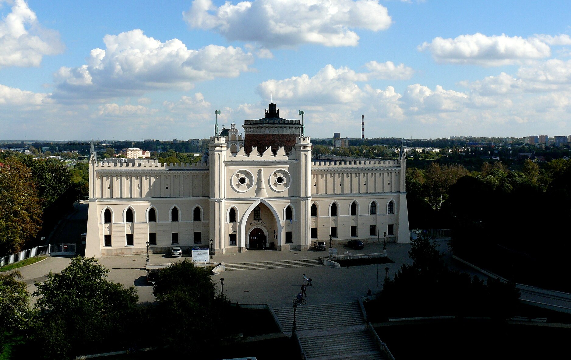 Zdjęcie przedstawia ścianę frontową zamku w Lublinie. Zamek posiada trzy kondygnacje. Na dwóch niższych kondygnacjach znajduje się szereg okien o łukowatym sklepieniu. Wejście do zamku, które jest po środku, zdobi łukowate duże sklepienie i dwie wieże po bokach. Do bramy zamku prowadzą schody, a do zamku prowadzi szeroka aleja, wokół rosną drzewa. W tle niebo. 
