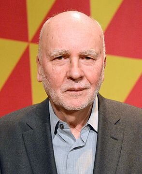 Zdjęcie przedstawia starszego mężczyznę. Jest niemal łysy. Ma gęste, siwe brwi i wydatny nos. Ma krótko ostrzyżoną brodę i wąsy. Ubrany jest w marynarkę i koszulę.