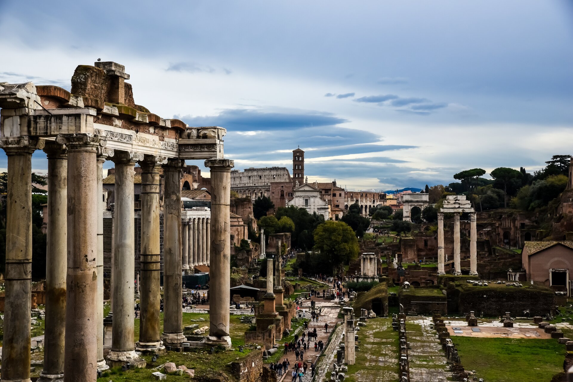 Ilustracja przedstawia aktualny stan Forum Romanum. Jest to kolorowa fotografia, która ukazuje górne fragmenty zachowanych kolumn. Po lewej stronie ilustracji znajduje się architraw świątyni podparty kolumnami z głowicami jońskimi. W tle, wśród drzew, widnieją ruiny pozostałych zabudowań. 