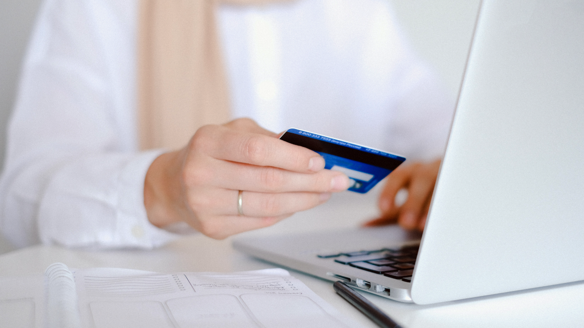 Zdjęcie przedstawia zbliżenie na ręce osoby, która siedzi przed komputerem, a w prawym ręku trzyma kartę płatniczą.