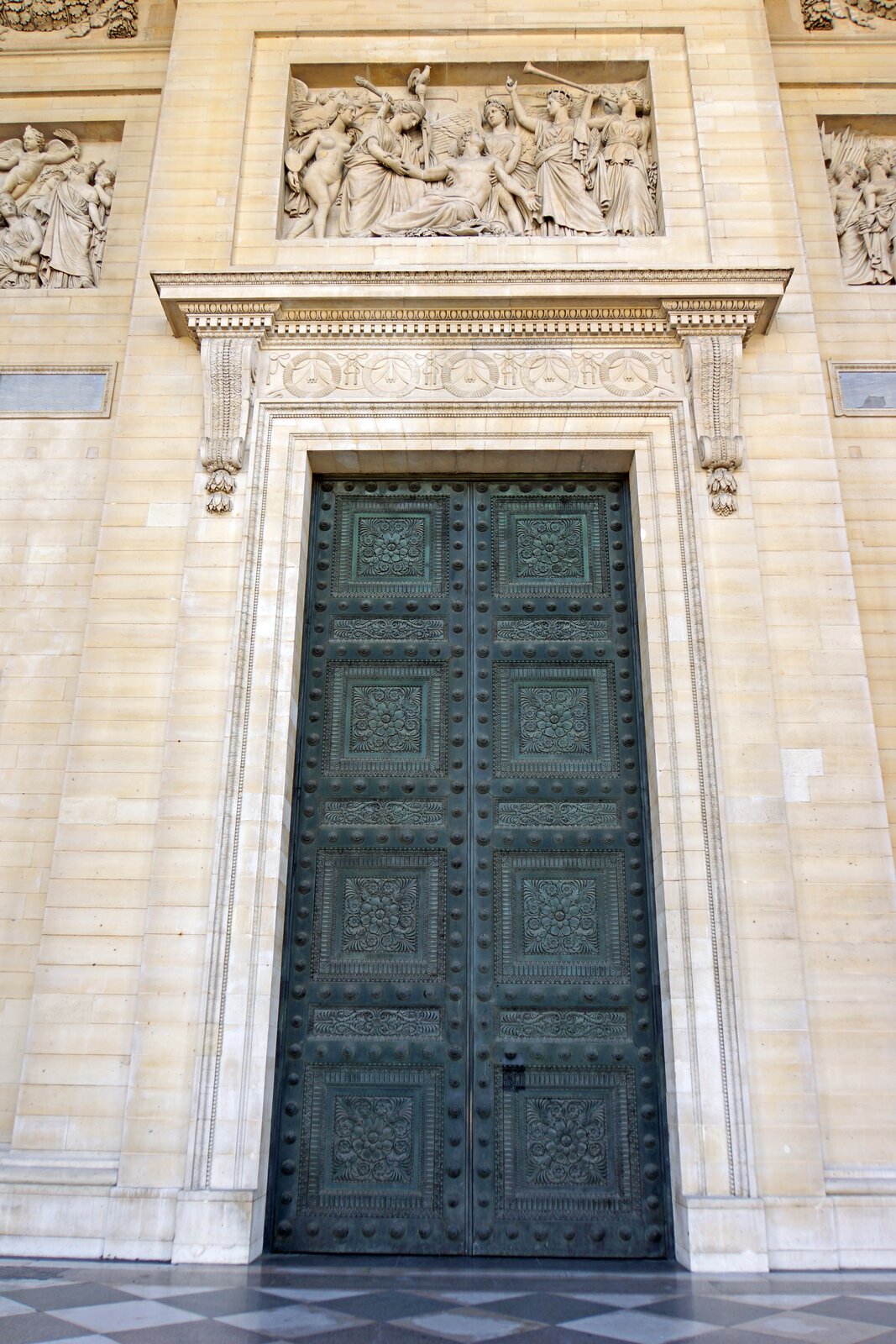 Zdjęcie przedstawia wysokie, metalowe drzwi, pokryte zieloną patyną. Mają one kwadratowe elementy, w których umieszczono kwiatowe zdobienia. Nad drzwiami znajduje się poziomy, wystający poza lico ściany element z żółtego kamienia, podobnie jak ściana. Nad nim umieszczono grupę rzeźbiarską w prostokątnej wnęce w ścianie. Rzeźby przedstawiają różne postacie. Jedna z nich dmie w trąbę. Jedna postać siedzi na ziemi, a inne ją podtrzymują.