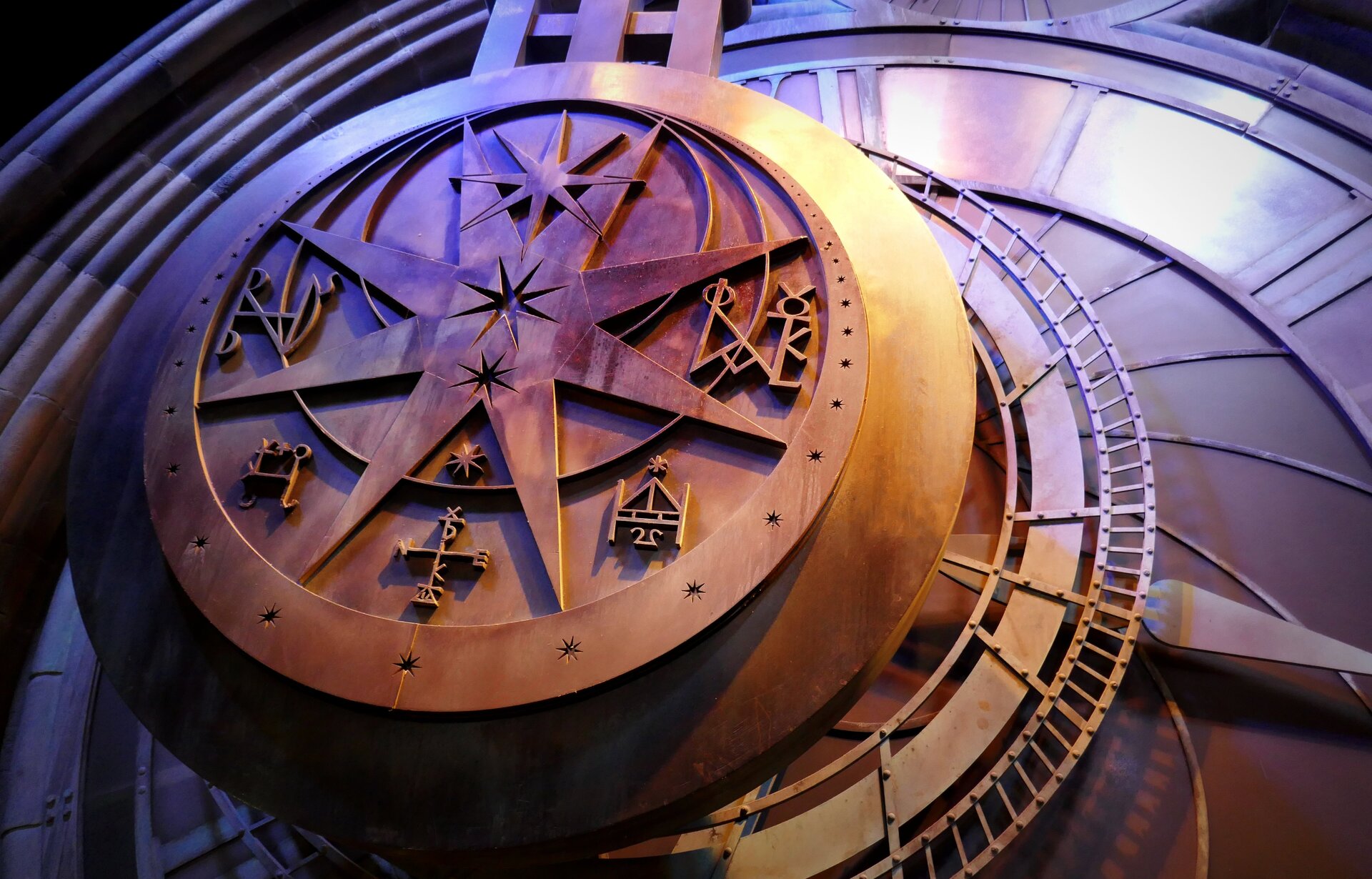 Fotografia przedstawia wahadło zabytkowego zegara z wieży kościelnej zdobione symbolami astronomicznymi. Na tle odchylonego wahadła dwie tarcze zegarowe: większa wskazuje godziny i fazy księżyca oraz mniejsza minutowa umieszczona w górnej części. Dookoła tarczy murowane wykończenie.