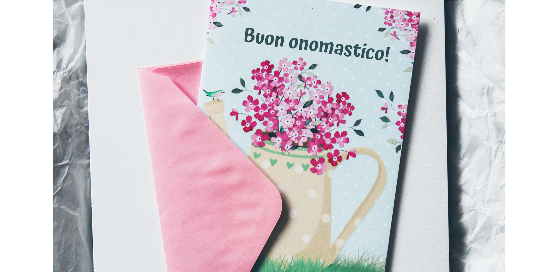 Na zdjęciu znajduje się kartka leżąca na kopercie. Na kartce widoczna jest konewka, a w niej bukiet kwiatów, Na konewce siedzi mały ptaszek. Znajduje się również napis „Buon obomastico!”. 