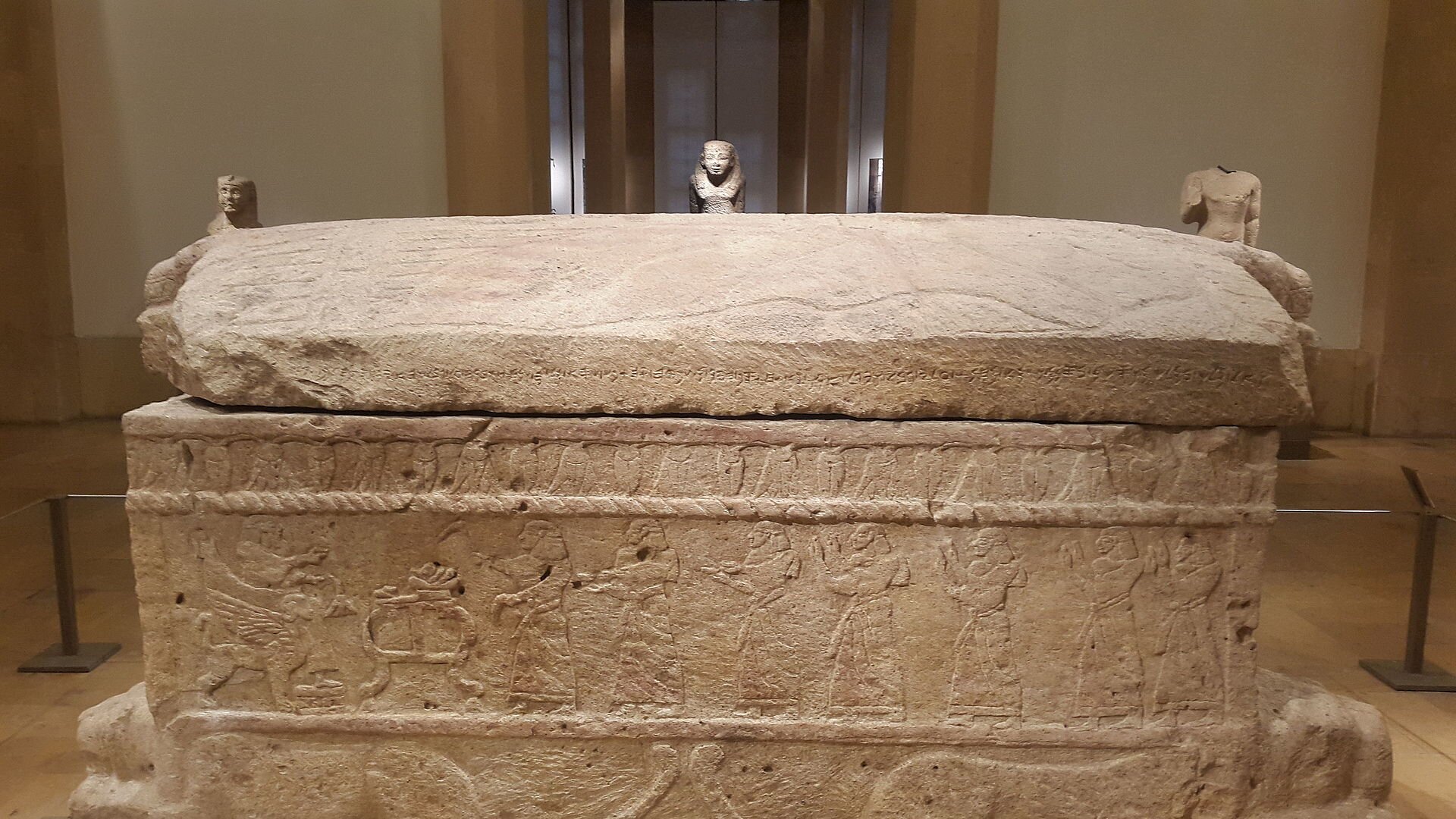 Zdjęcie przedstawia kamienny grobowiec, w kształcie poziomego, leżącego prostopadłościanu. Sarkofag spoczywa na postaciach leżących lwów, których ciała przedstawiono w reliefie na dłuższych bokach, podczas gdy głowy w pełnej rzeźbie (po dwie z każdej strony) wystają na krótszych. Na każdej ze ścian sarkofagu przedstawione są sceny figuralne, nad którymi biegnie ciągły fryz pączków i kwiatów lotosu zwieszonych w dół.