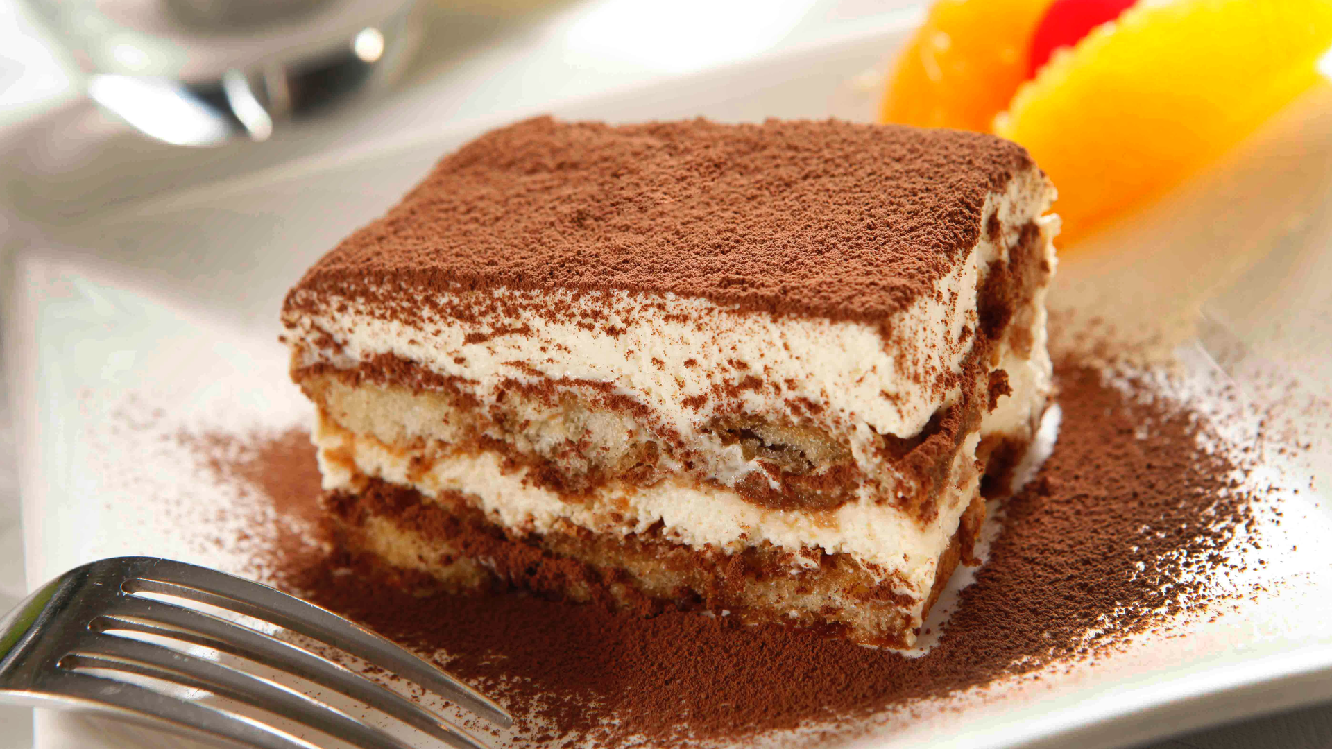 Zdjęcie przedstawia bardzo ładnie podany deser Tiramisù na porcelanowym, kwadratowym talerzyku. Widoczna jest każda warstwa deseru, czyli biszkopty przełożone masą serową. Tiramisù  na wierzchu posypane jest kakao. 