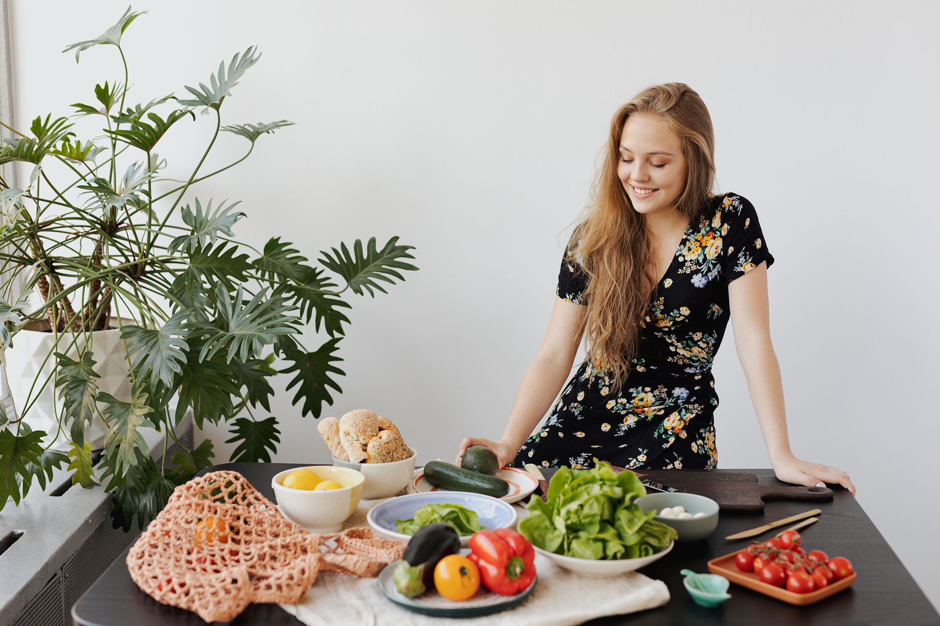Zdjęcie przedstawia młodą kobietę stojącą przy stole, na którym są rozłożone warzywa. Są tam: pomidorki cherry sałata, papryka, żółty pomidor, bakłażan, cukinia i awokado. Część warzyw leży w plecionej siatce na zakupy. 