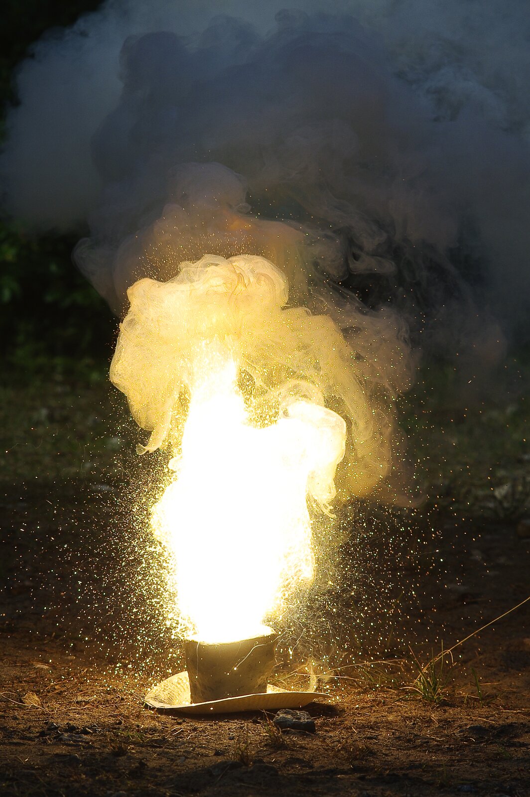 Zdjęcie przedstawiające żółty płomień wydobywający się z pojemnika, który stoi na podstawce. Nad płomieniem są kłęby jasnego dymu. Wokół ognia spora ilość iskier.