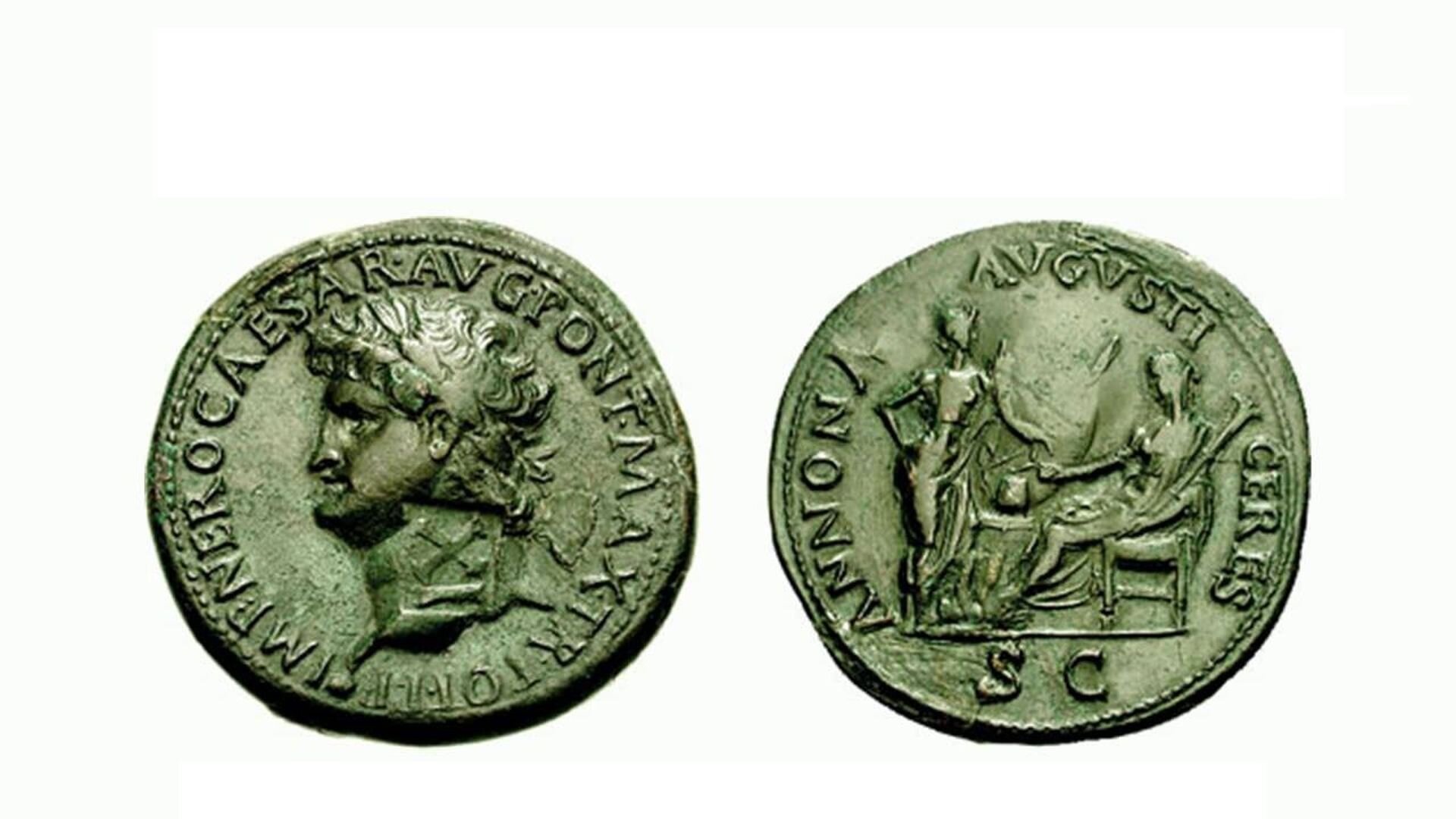 Ilustracja przedstawia widok na starożytną monetę, która została wykuta za czasów Nerona. Moneta ma barwę zieloną, prawdopodobnie od patyny. Na awersie znajduje się twarz Nerona, na rewersie dwie postacie.