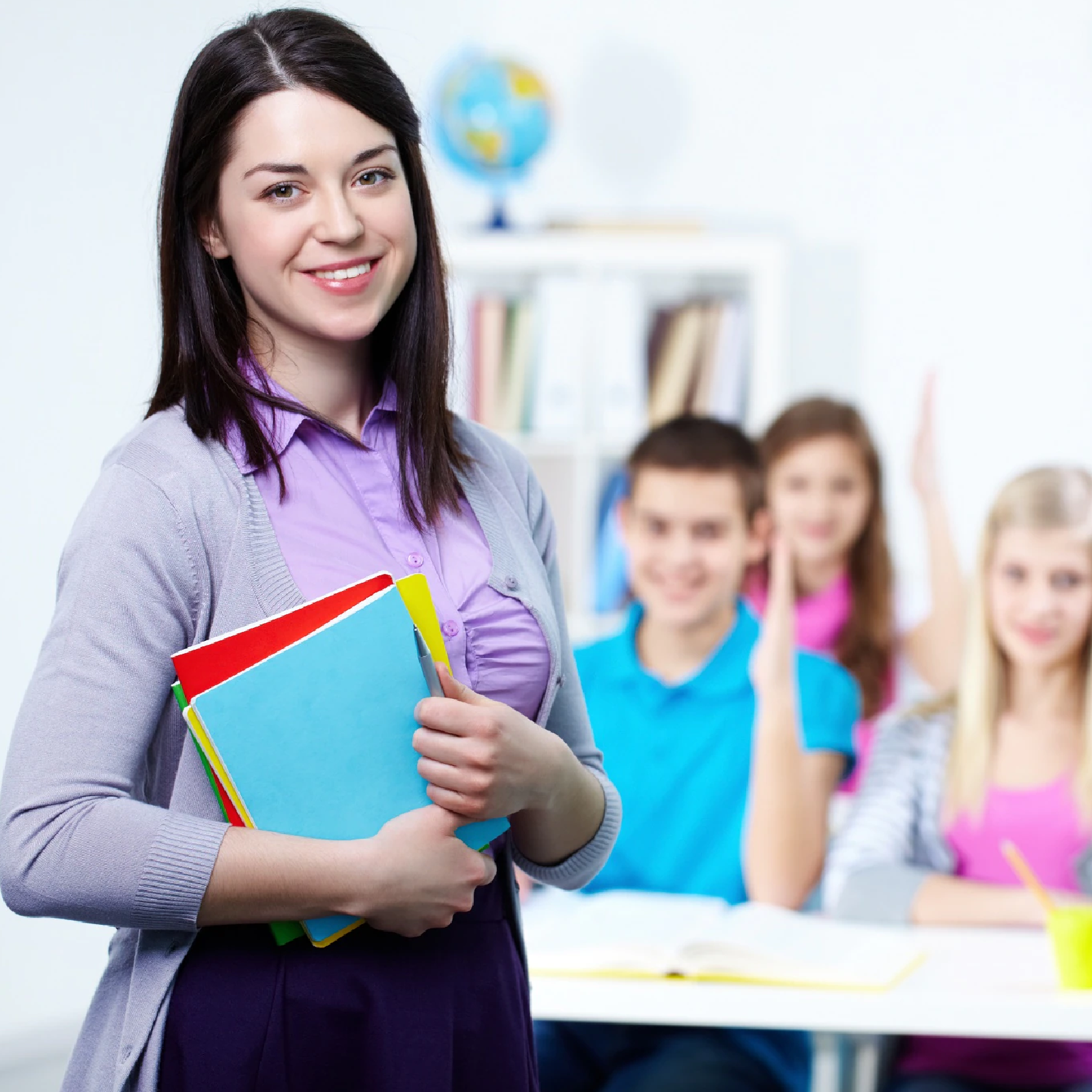 Ilustracja przedstawia nauczycielkę trzymającą kolorowe zeszyty. W tle widoczni są uczniowie zgłaszający się poprzez podniesienie ręki.