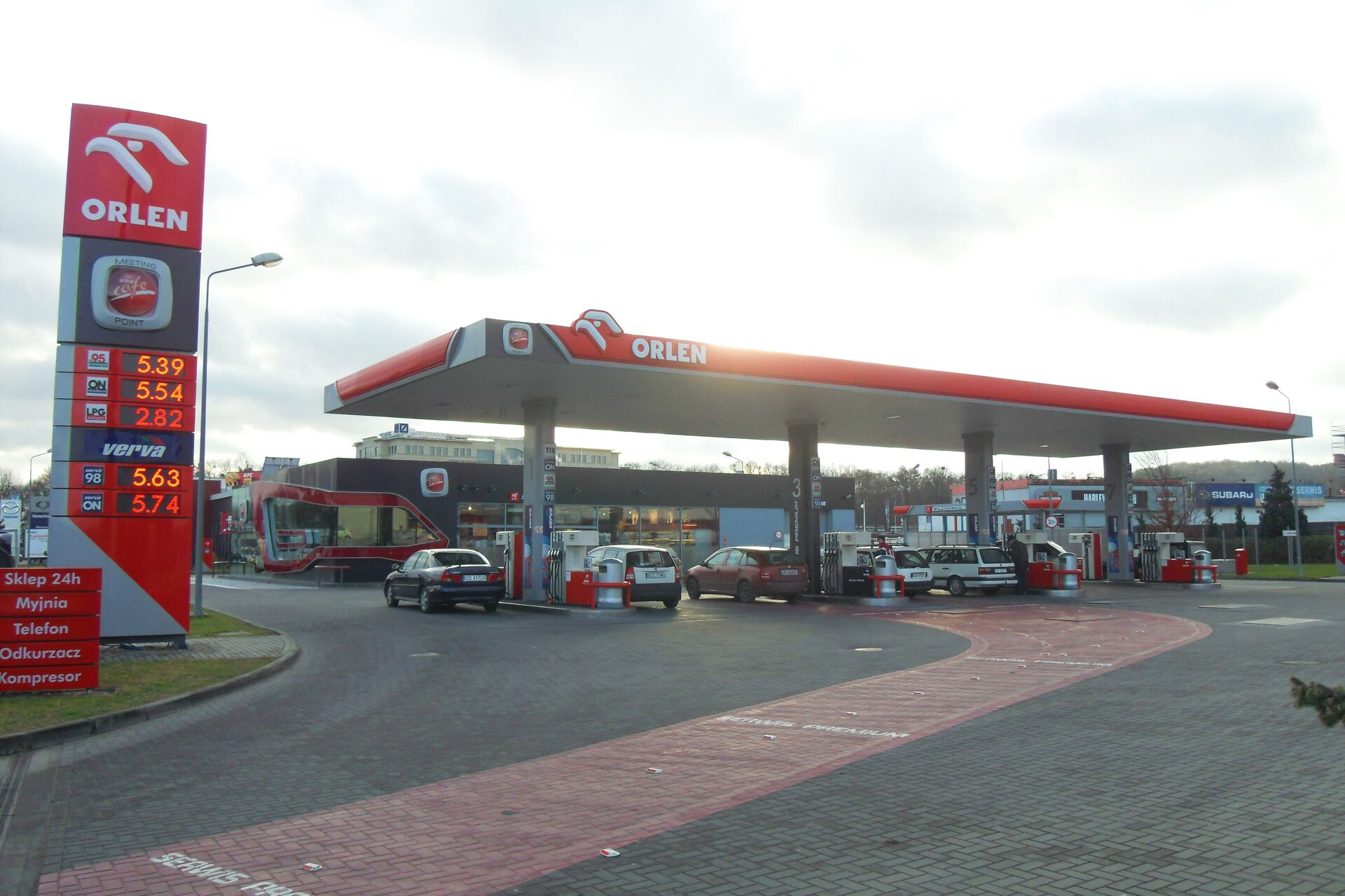 Zdjęcie przedstawia stację benzynową ORLEN. Przy dystrybutorach stoją samochody. Przy wjeździe stoi wysoka tablica z cenami poszczególnych paliw.