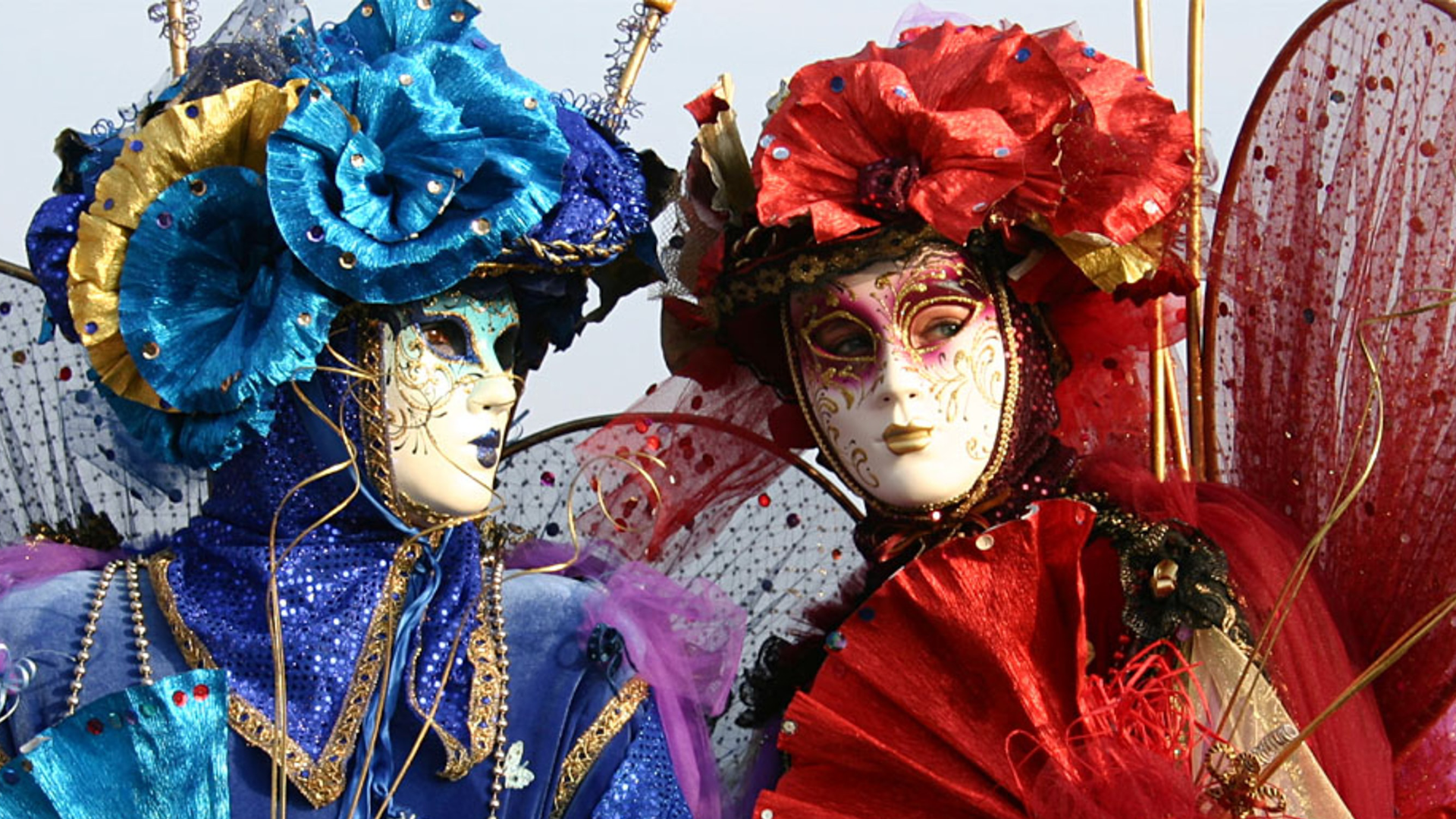 Zdjęcie przedstawia dwie kobiety ubrane w kolorowe stroje  i nakrycia głowy. Na twarzach mają maski weneckie.