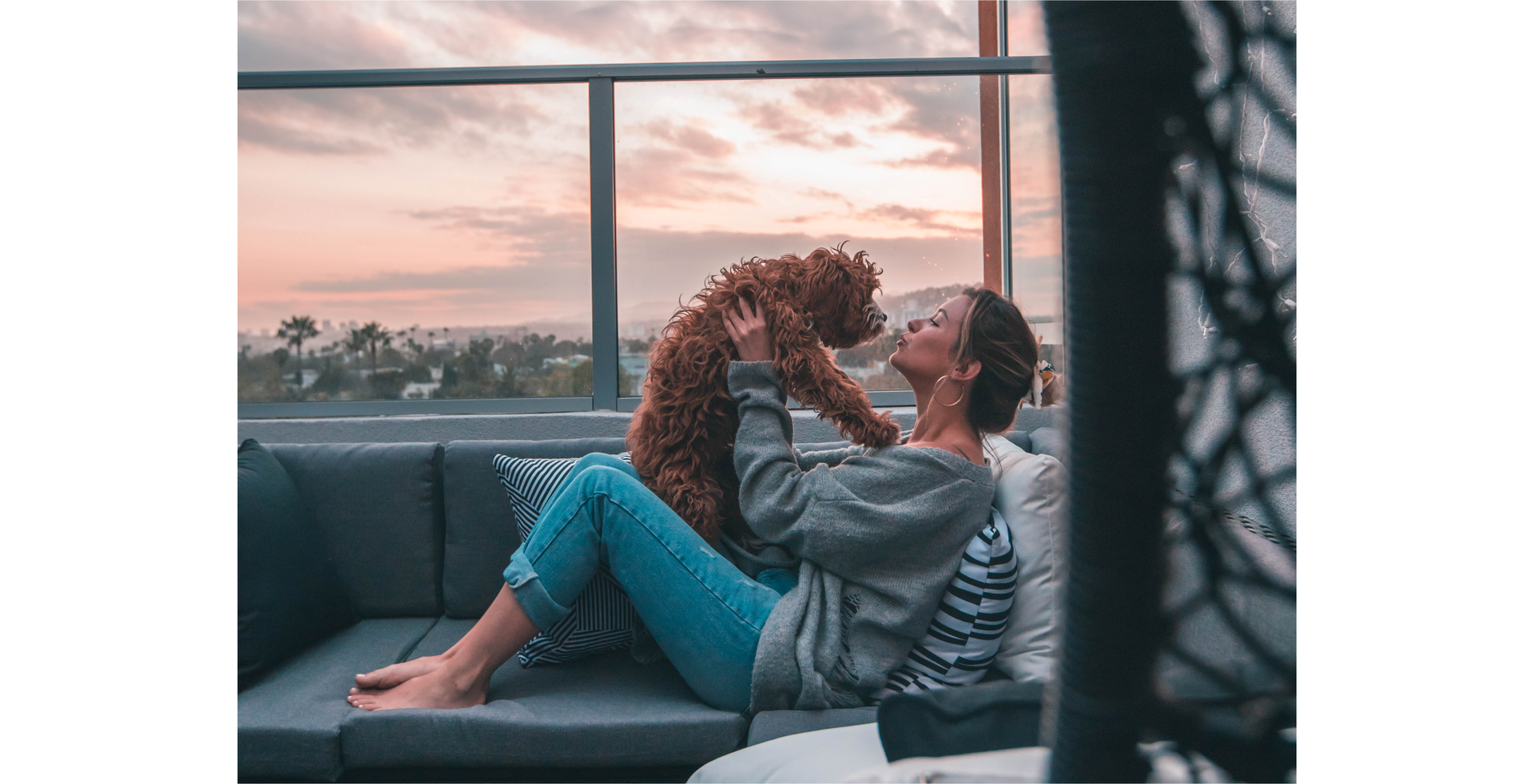 Zdjęcie przedstawia kobietę siedzącą na kanapie przy dużym oknie. Na kobiecie siedzi pies. Kobieta zbliża pysk psa do swojej twarzy. Na drugim planie widoczne jest niebo o zachodzie słońca.