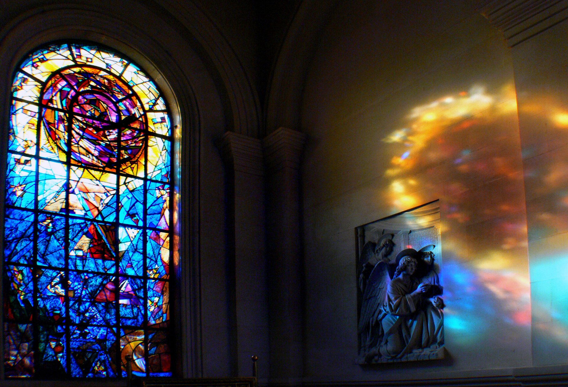 Ilustracja przedstawia witraż w kościele w Christchurch w Nowej Zelandii,. Ukazuje fotografię z widokiem na półokrągłe okno wypełnione w dolnej części niebieskimi szkiełkami, a w górnej okręgiem w ciepłych barwach. Kolorowe światło odbija się na ścianie z płaskorzeźbą.