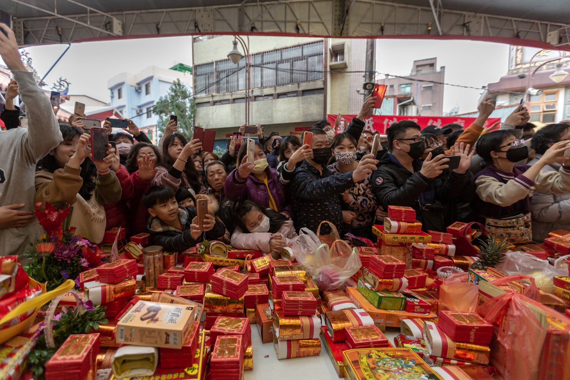 Zorientowana poziomo fotografia barwna przedstawiająca zgromadzony za straganem targowym trzymających w podniesionych dłoniach telefony komórkowe typu smartfon. Na pierwszym planie blat straganu zasłany towarem zapakowanym w nieduże, czerwone pudełka. Poniżej górnej krawędzi fotografii widoczny fragment metalowego dachu straganu. W tle wysoki, biały budynek otoczony mniejszymi domami.