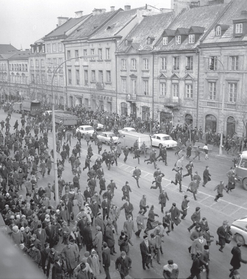 Ilustracja przedstawia ulicę, wzdłuż której stoją kamienice. Na ulicy tłum biegnących, rozpierzchających się ludzi. Wśród tłumu są cywile oraz umundurowani funkcjonariusze.
