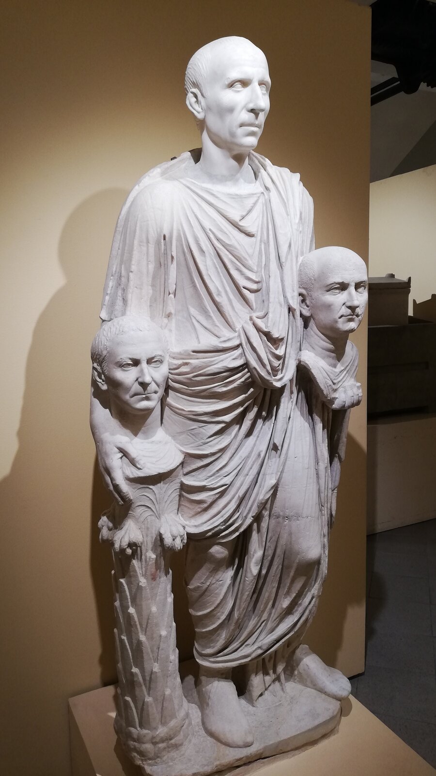 Ilustracja przedstawia „Statuę Barberini”, anonimowe dzieło rzymskie datowane na 40–30 p.n.e., przechowywane w Muzeach Kapitolińskich w Rzymie. Ukazuje stojącą postać mężczyzny ubranego w obszerną togę, której liczne fałdy opadają pionowo wzdłuż ciała. Figura trzyma w obu dłoniach dwa marmurowe popiersia ustawione na niskich, cylindrycznych podporach przypominających fragmenty pni lub kolumienek.
Centralna postać ma poważny, realistycznie opracowany wyraz twarzy. Oba trzymane popiersia również przedstawiają mężczyzn, każdy z indywidualnie oddanymi rysami. Całość wykonana jest z jasnego marmuru, a kompozycja ma charakter frontalny i statyczny.