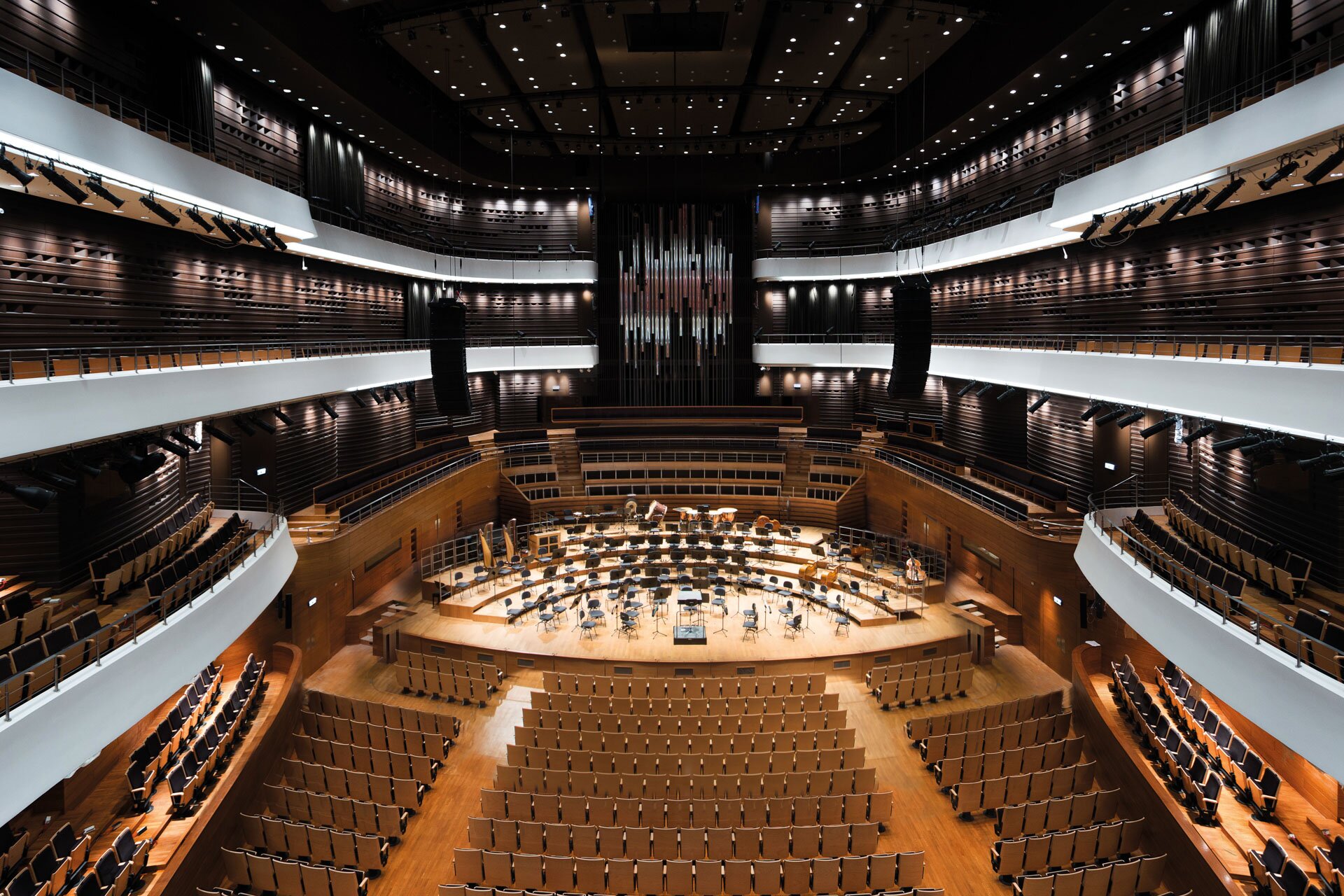 Fotografia przedstawia dużą salę koncertową. Centralną część stanowi scena, na niej stoją krzesła i stojaki na nuty oraz pulpit dyrygenta. Przed sceną fotele widowni. Sala jest otoczona balkonami w białym kolorze, znajdują się one na trzech poziomach. Sala jest utrzymana w różnych odcieniach brązu.