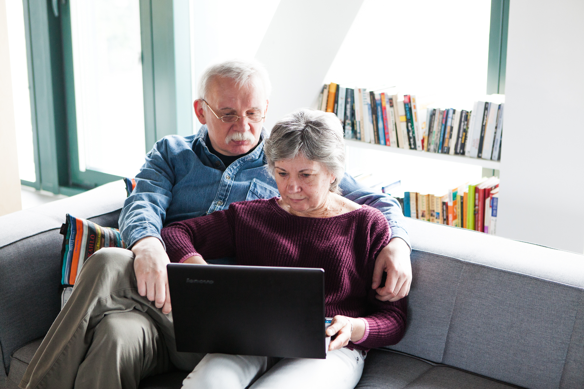 Fotografia przedstawia parę ludzi w wieku około 65 lat, siedzących razem na kanapie przy komputerze. Za nimi półki z książkami.