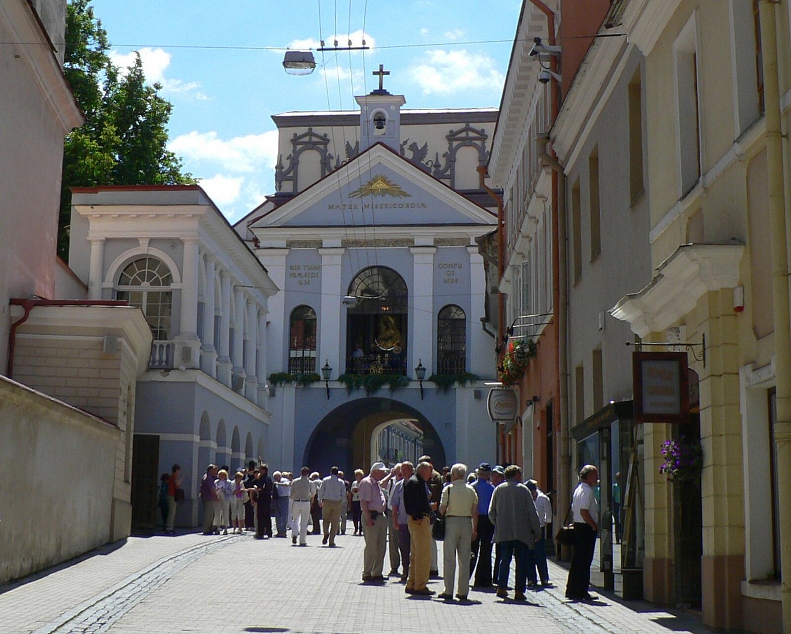 Fotografia przedstawia Ostrą Bramę w Wilnie. Głównym elementem bramy jest błękitny budynek, w którego podstawie znajduje się łuk, przez który przechodzą ludzie. Nad łukiem okno, za którym znajduje się kapliczka z obrazem Matki Boskiej. Obraz widoczny również z poziomu ulicy.
