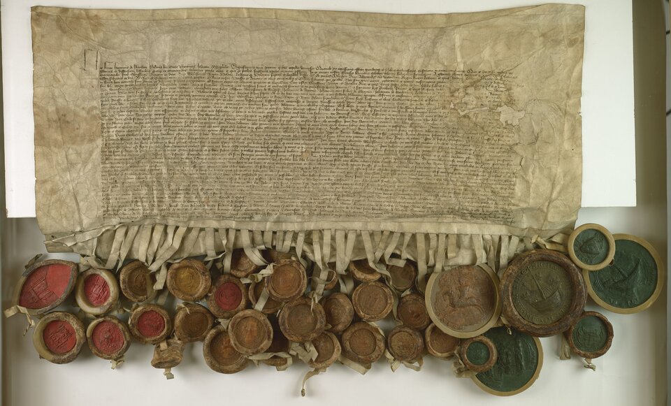 Zdjęcie przedstawia dokument pierwszego pokoju toruńskiego. Na pergaminie jest gęsto zapisana treść traktatu. Na dole jest on pocięty na pionowe paski, do których przypięte są pieczęcie. Dokument leży na białym stole.